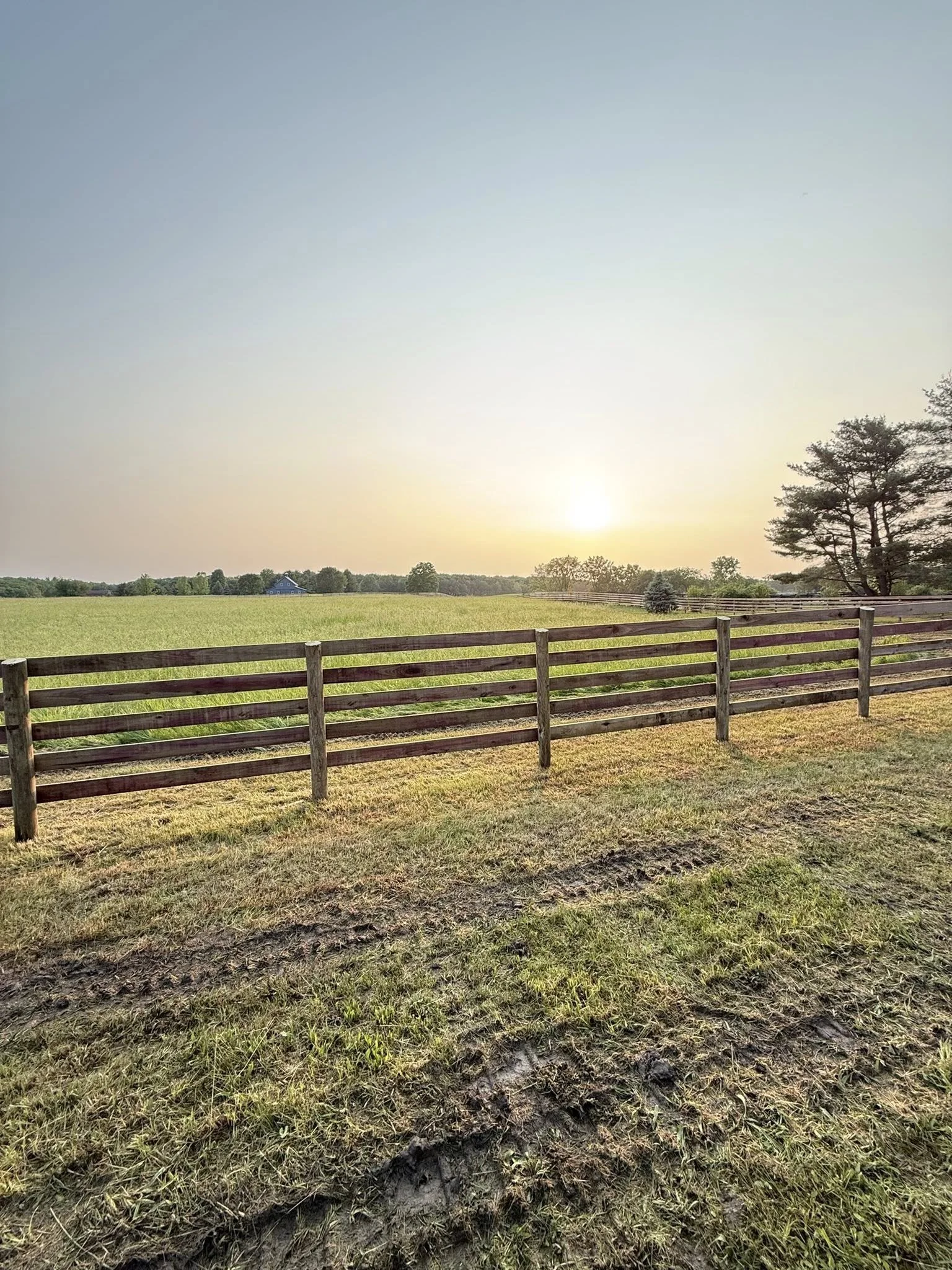 A scenic view of a grassy field with a wooden fence in the foreground, trees to the right, a small house in the distance, and the sun setting or rising on the horizon under a clear sky.