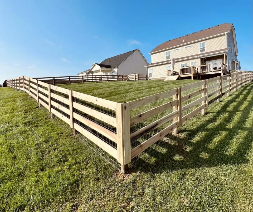 New wooden fence surrounding a grassy backyard of a house with a deck, on a sunny day.