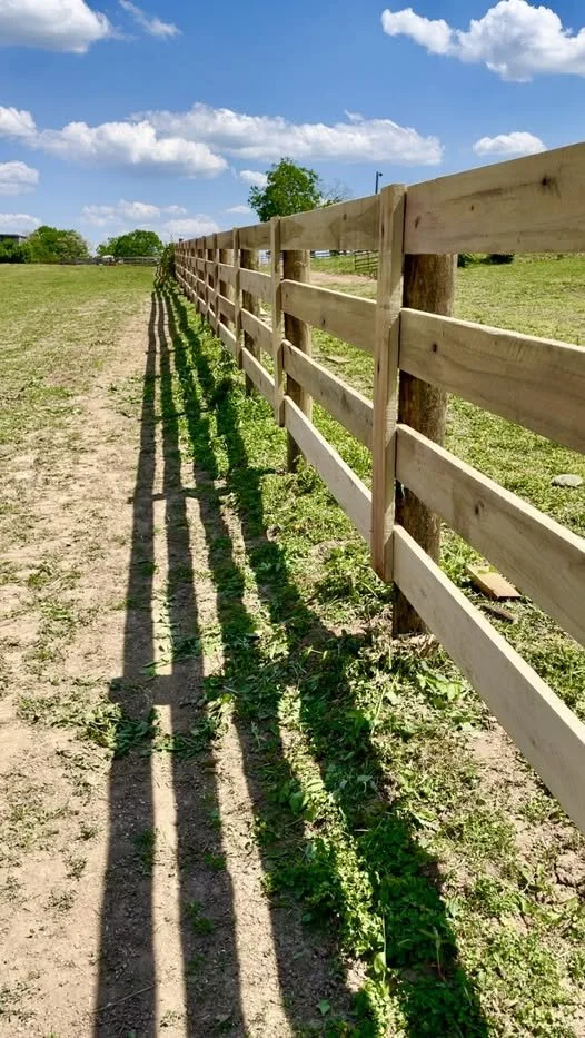 Wooden fence casting shadows on a dirt path in a grassy field under a blue sky with white clouds.