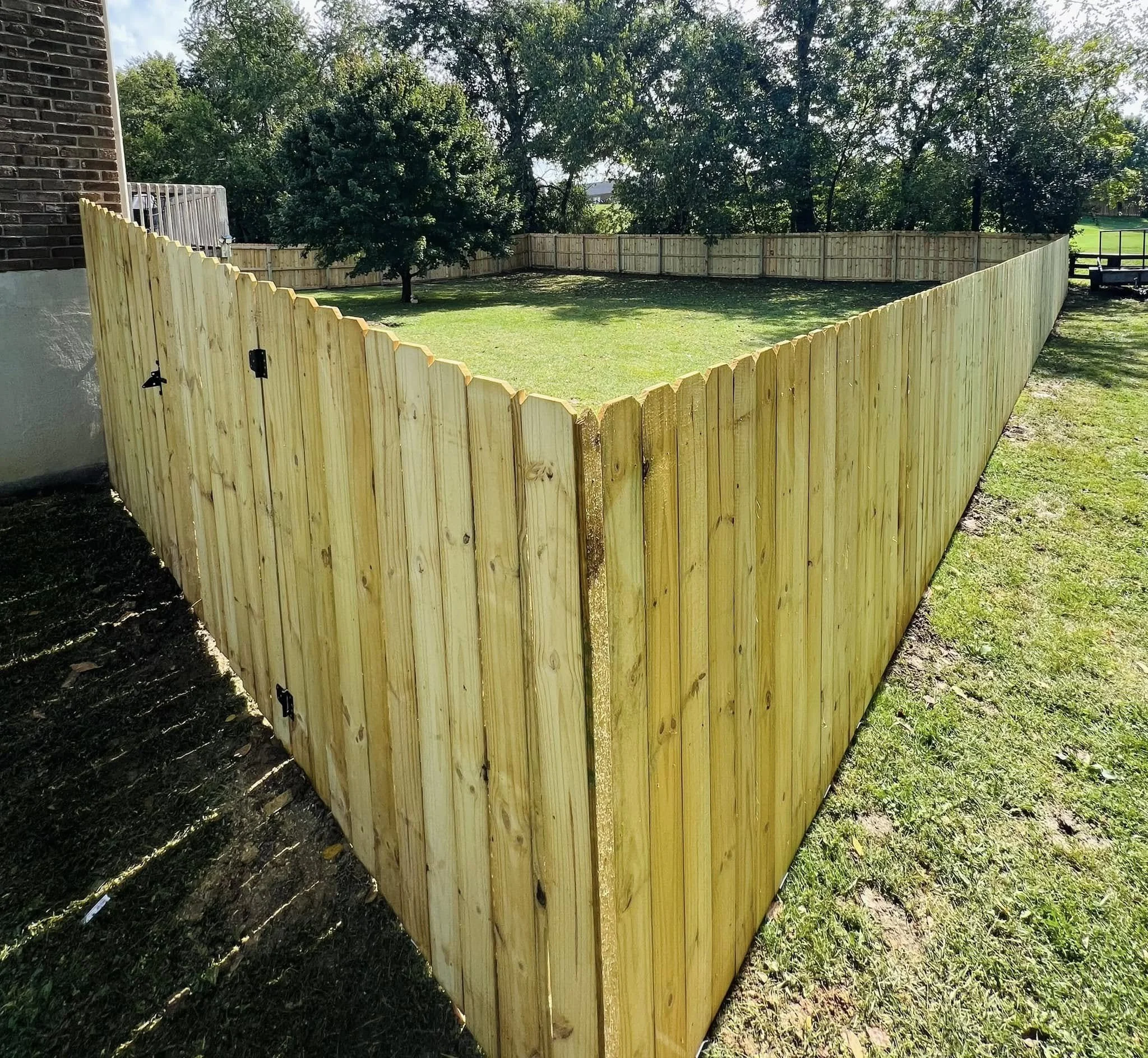 A wooden fence surrounding a grassy backyard with trees and a clear blue sky.