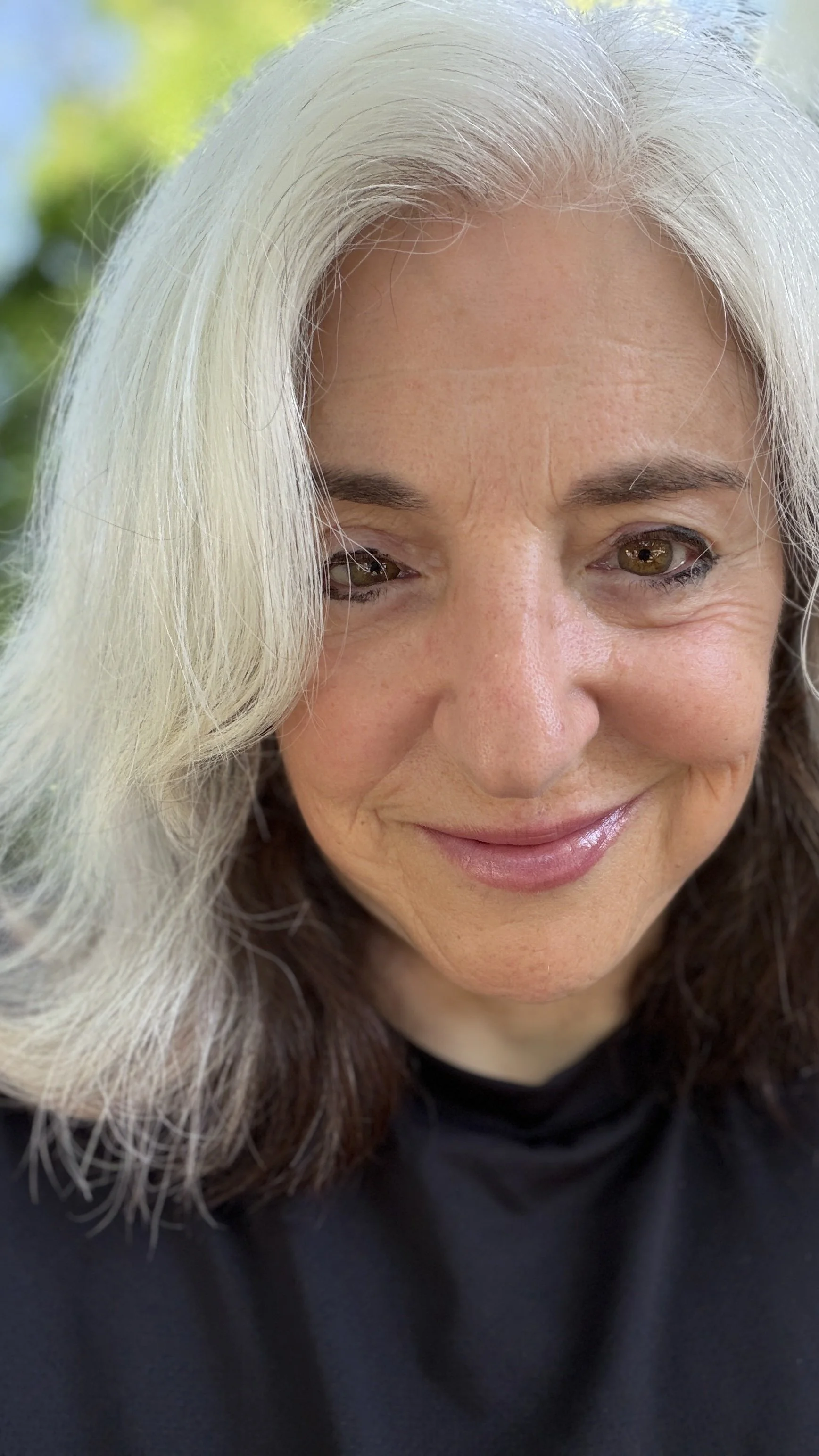 Close-up of a smiling woman with long gray and brown hair, outdoors with green foliage in the background.