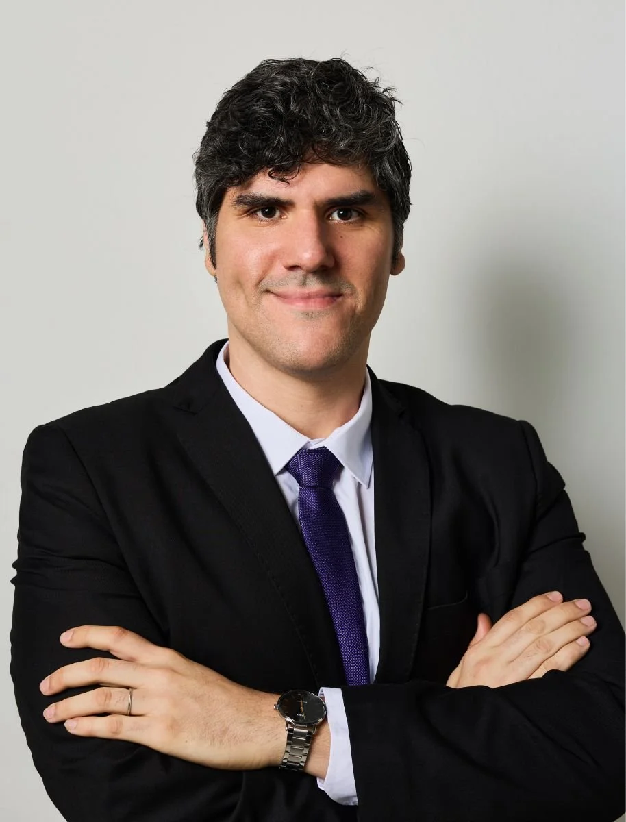 Professional man with dark, curly hair, wearing a black suit, white shirt, purple tie, and a watch, standing with arms crossed, smiling, against a plain light background.
