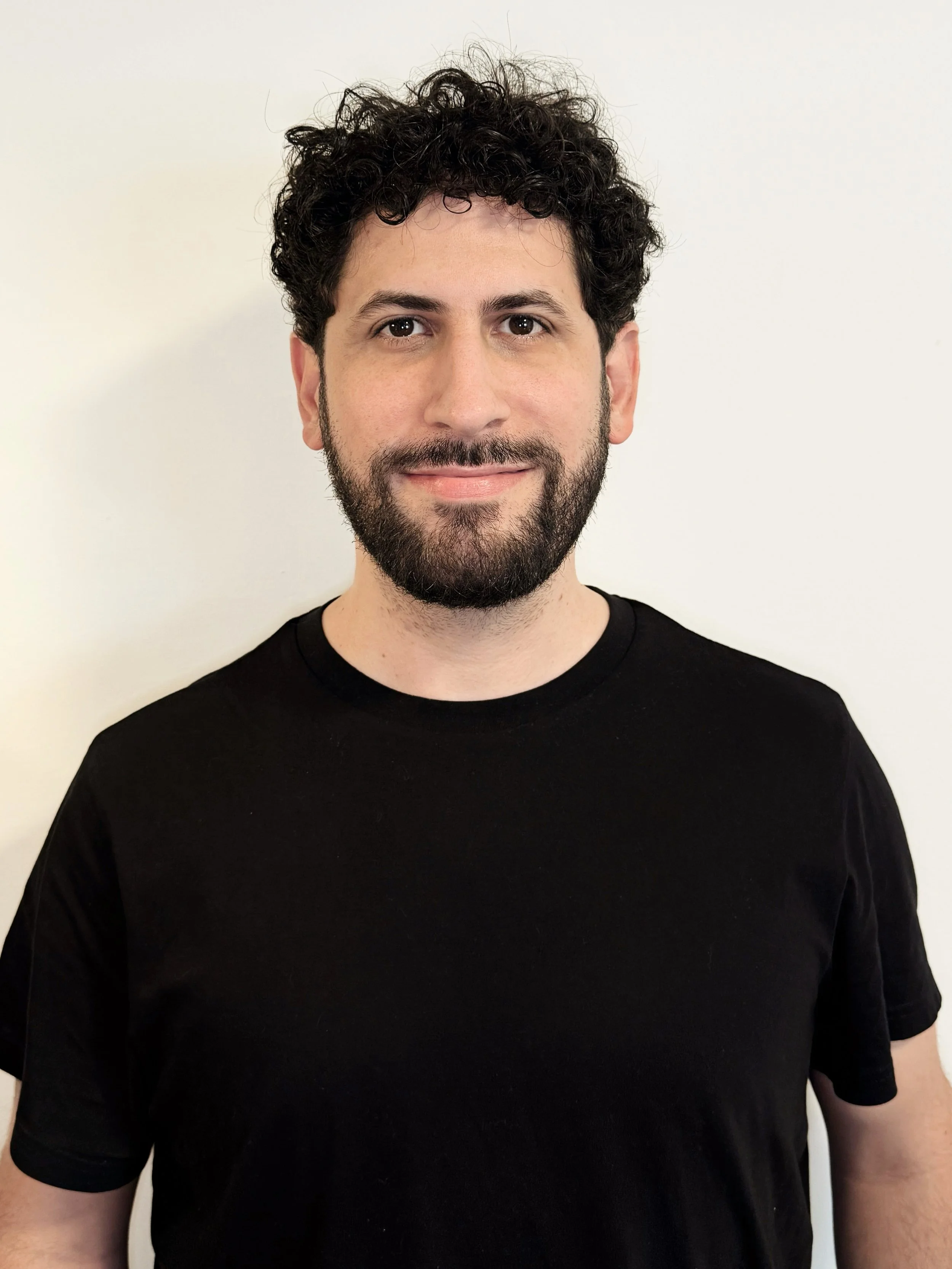 A man with curly dark hair, a beard, and fair skin, wearing a black t-shirt, standing against a plain white background.