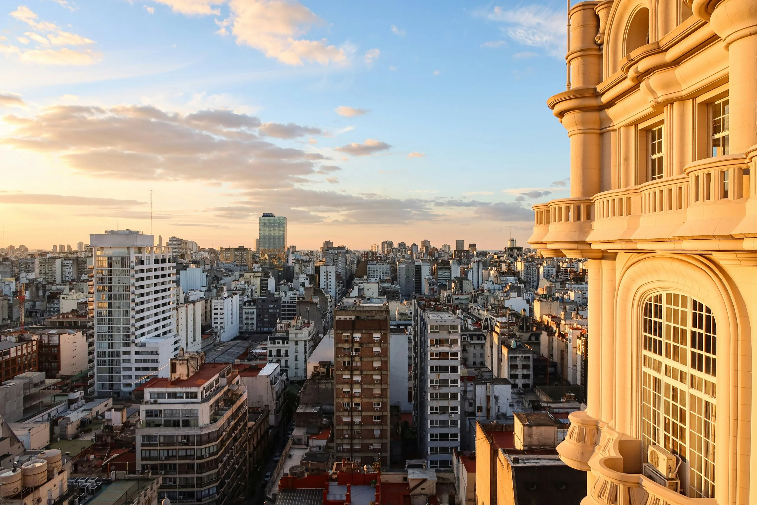 City skyline viewed at sunset with a tall ornate building on the right side and modern high-rise buildings in the distance, under a sky with scattered clouds.