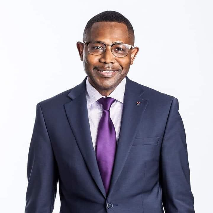 A professional man in a navy blue suit, white shirt, purple tie, wearing glasses, smiling, against a plain white background.