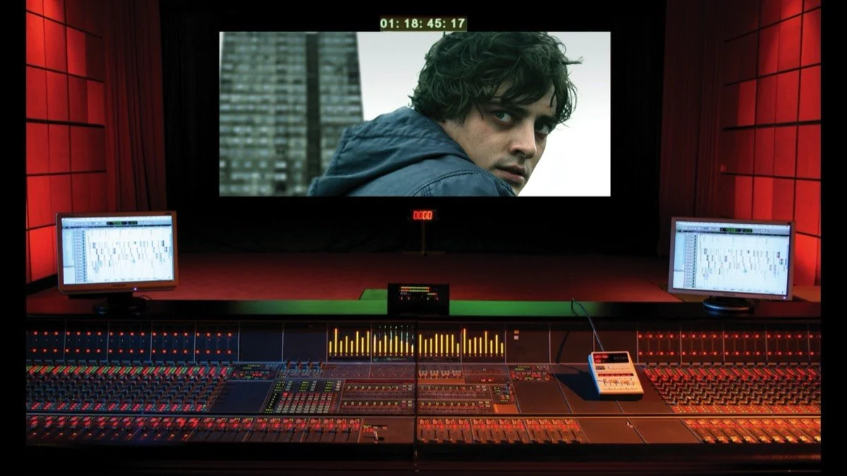 A sound editing studio with a large mixing console, two monitors displaying audio waveforms, and a big screen showing a close-up of a man with curly hair wearing a blue jacket.