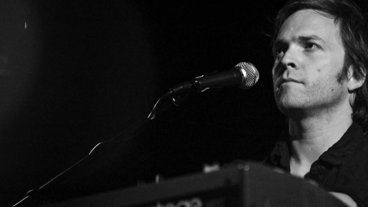 Black and white photo of a man with medium-length hair, looking thoughtfully at a microphone during a performance or recording session.