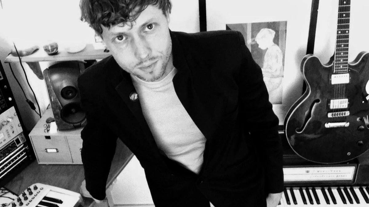 Black and white photo of a young man with curly hair, wearing a white t-shirt and dark jacket, standing in a room with musical instruments including a guitar, keyboard, and speakers.