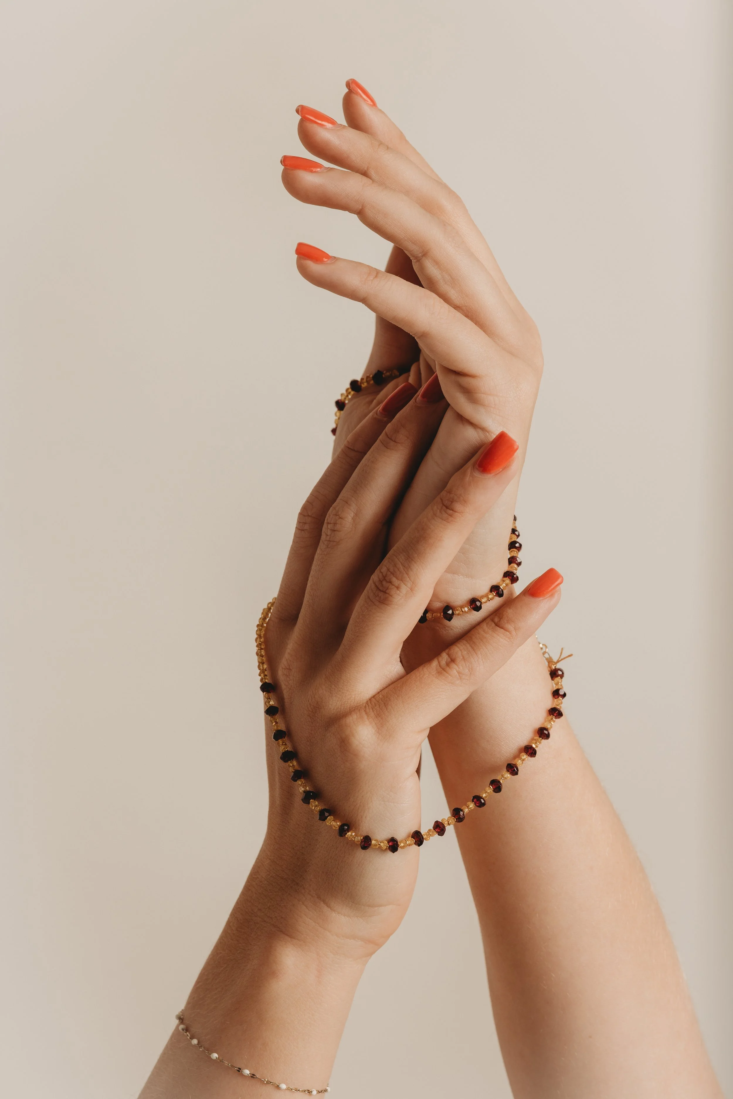 Two hands with manicured nails, one with a coral shade and the other with a natural tone, are intertwined against a light background. They are adorned with multiple beaded bracelets in black, red, and gold colors.