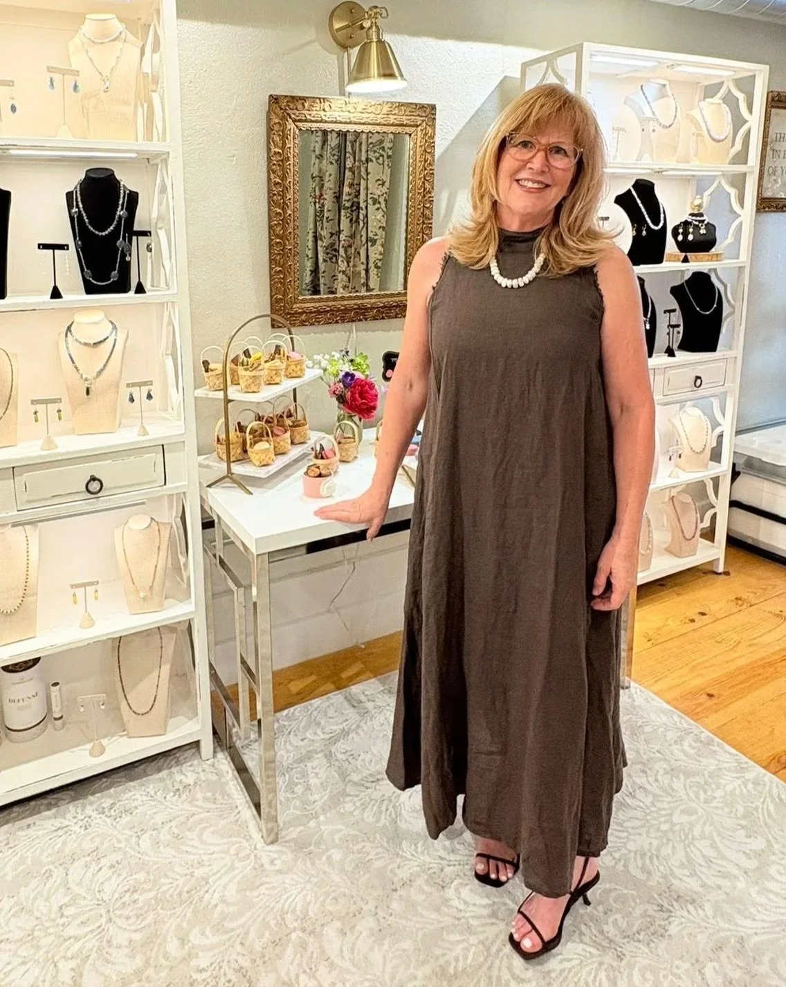 A woman standing in a jewelry store, wearing a sleeveless brown dress and black high heels, with jewelry displays and shelves of necklaces and accessories around her.