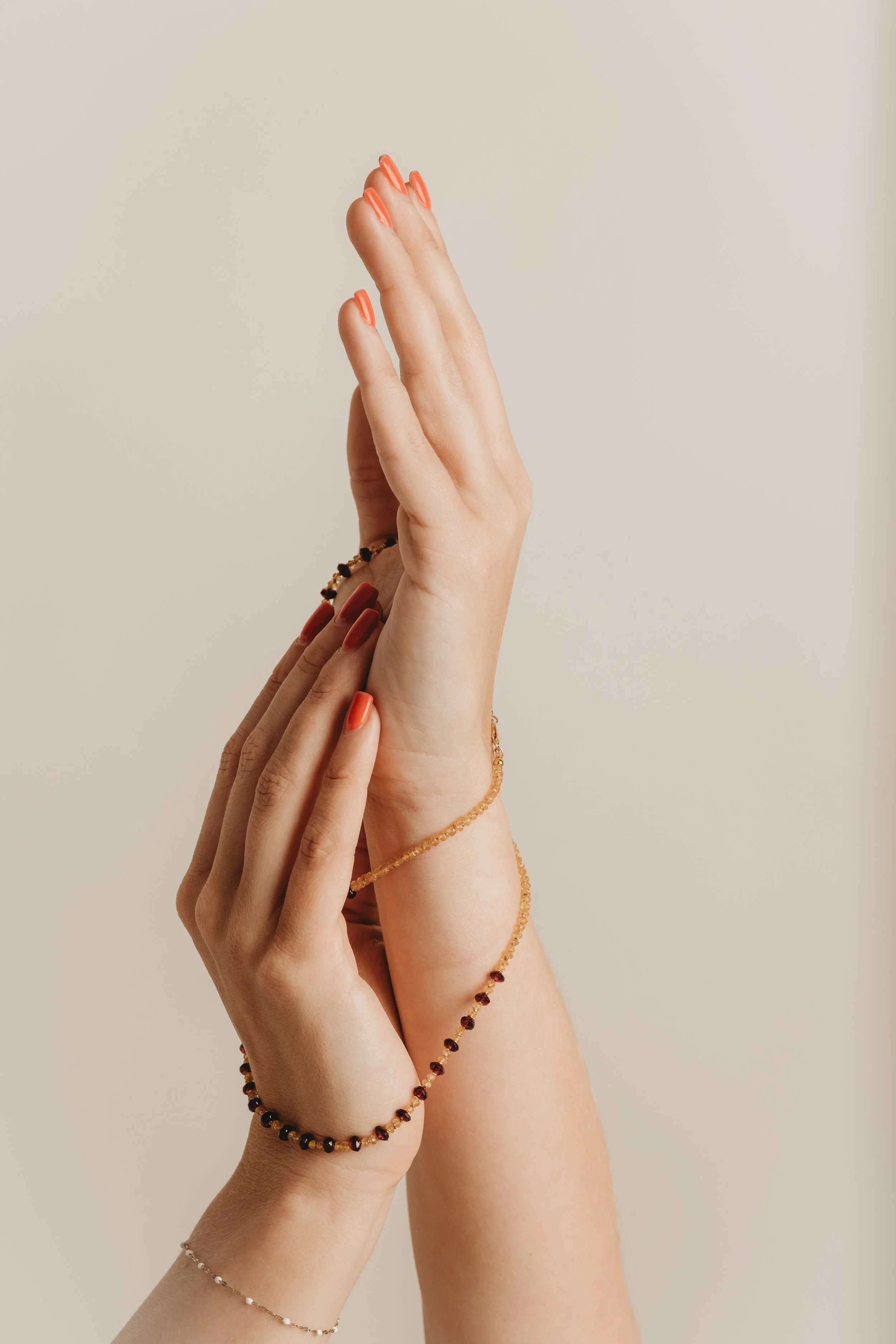 Close-up of hands with coral-colored nail polish and jewelry, including bracelets and rings, against a neutral background.