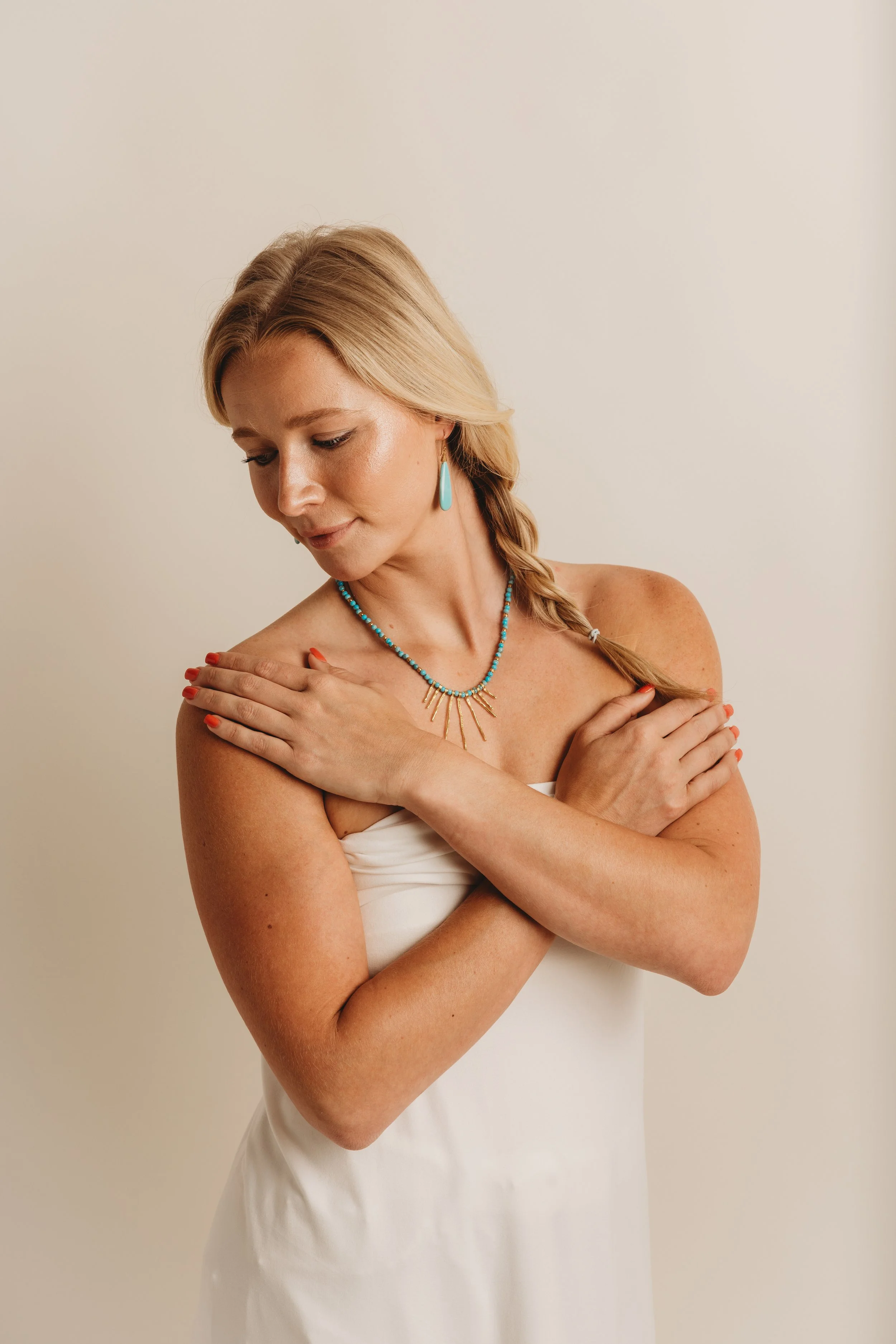 A woman with blonde hair in a braid, wearing turquoise jewelry, with arms crossed over her chest, against a plain light background.