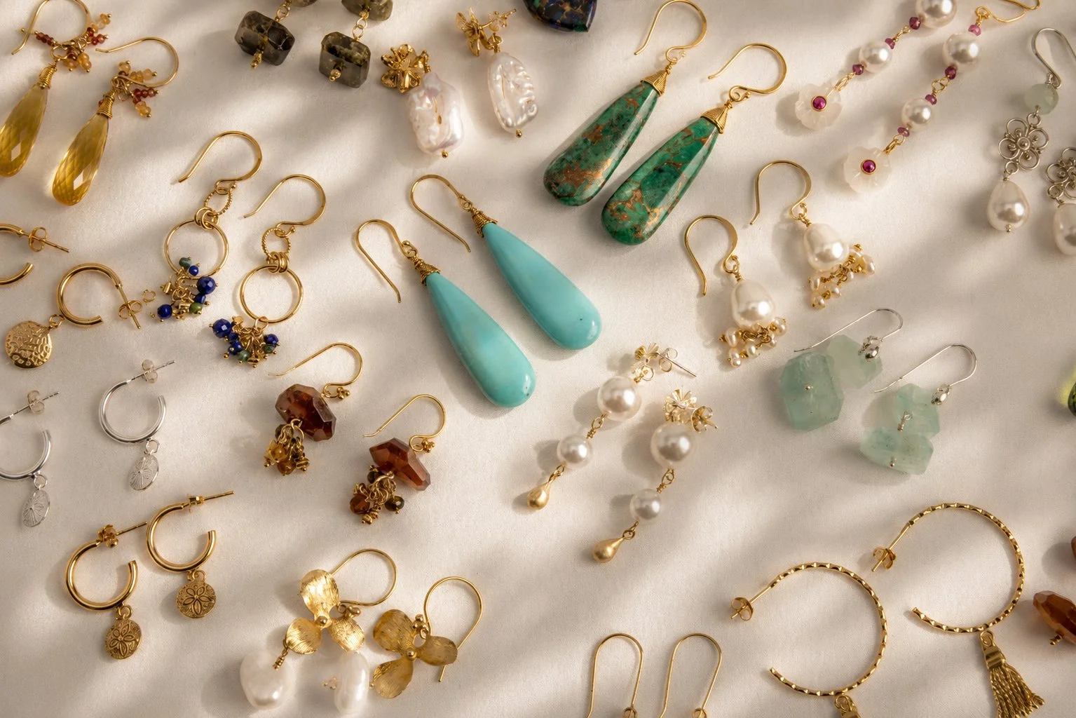 Various pairs of earrings displayed on a white surface, including gold hoops, drop earrings with turquoise and green stones, pearl earrings, and beaded designs.
