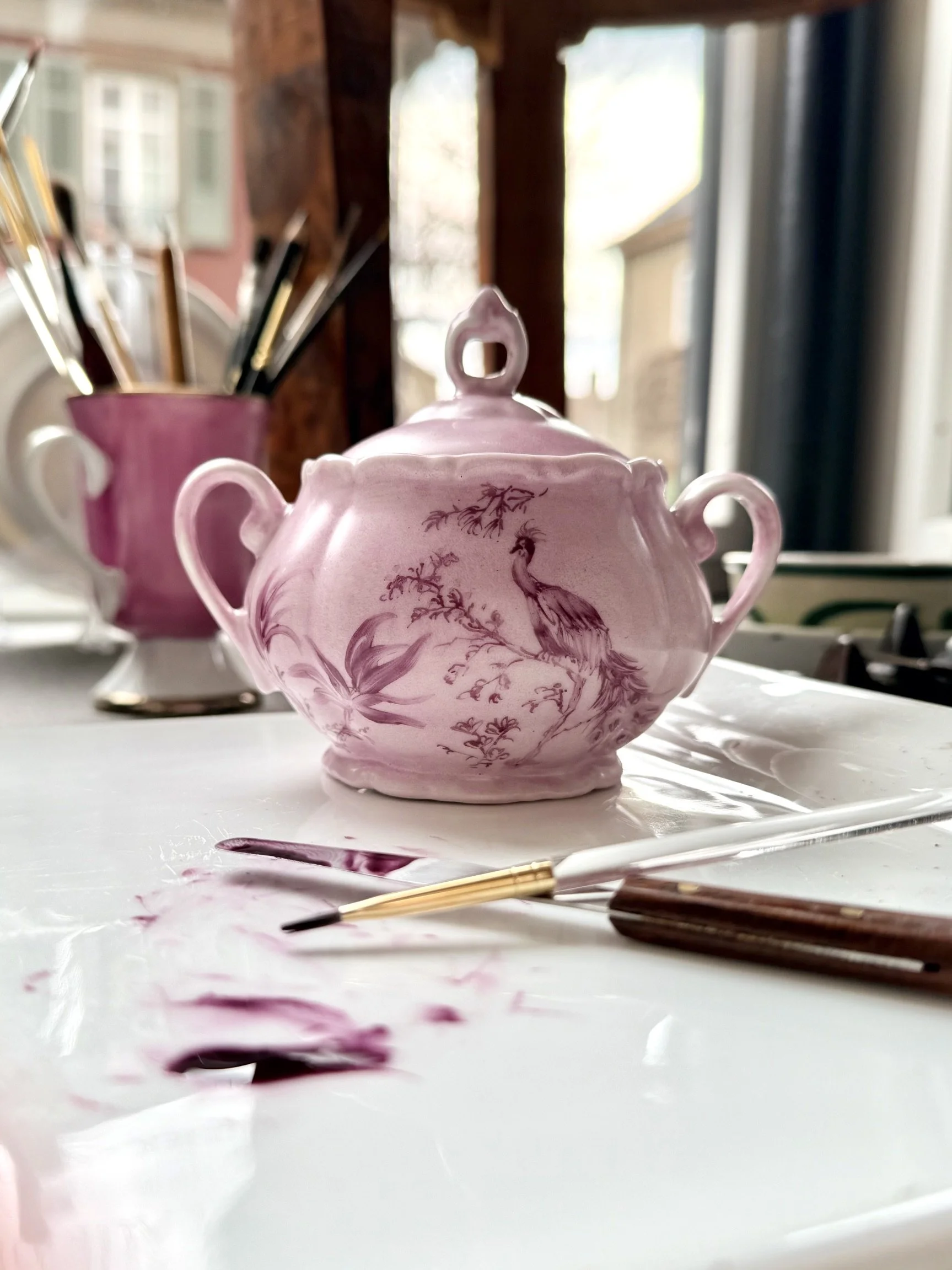 A pink porcelain teapot with painted peacocks and trees, placed on a white surface with art brushes and spilled pink paint in front of it, set near a window with sunlight.