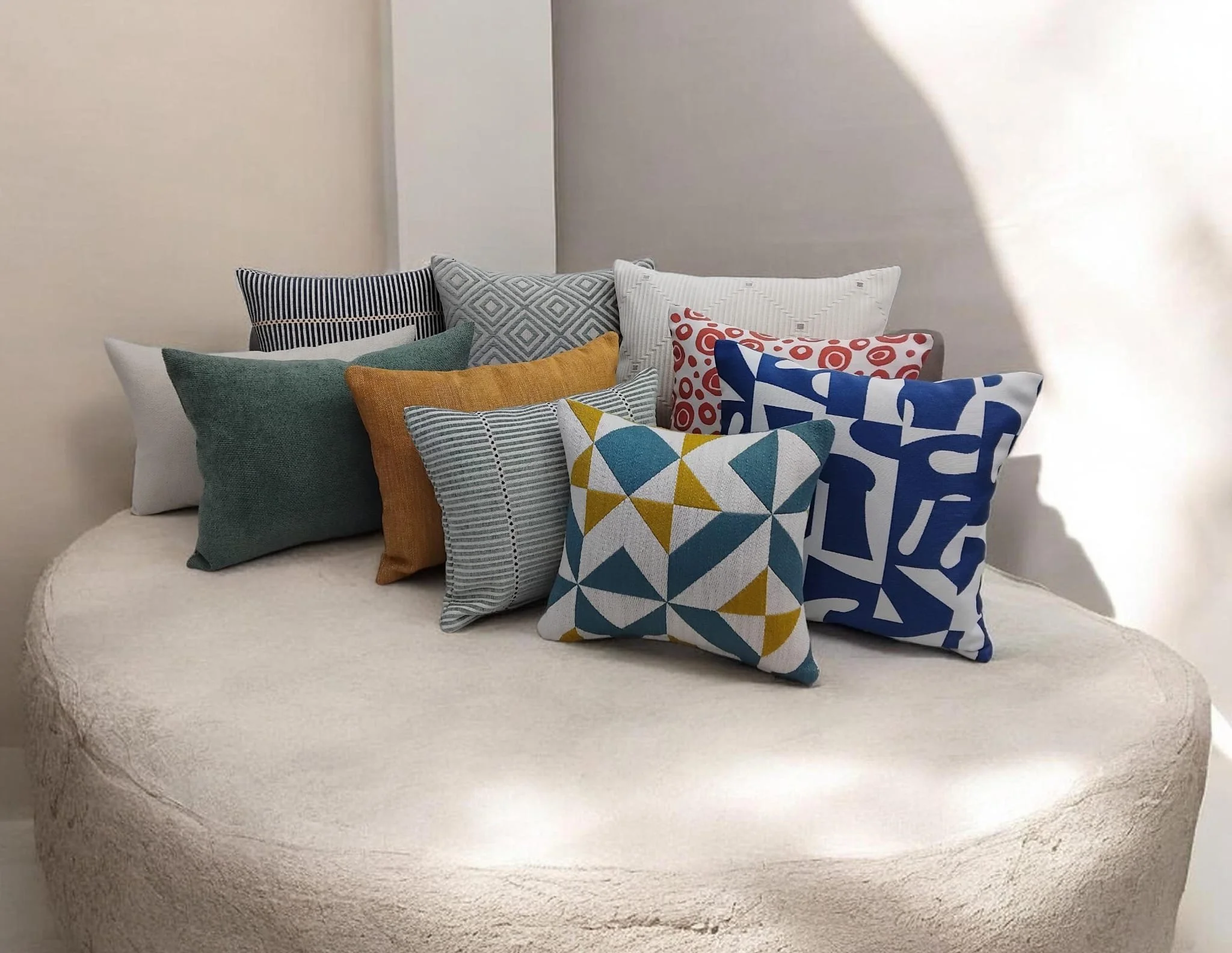 A white cushioned bench or sofa with a curved edge, decorated with multiple colorful patterned pillows, situated in a plain off-white room.