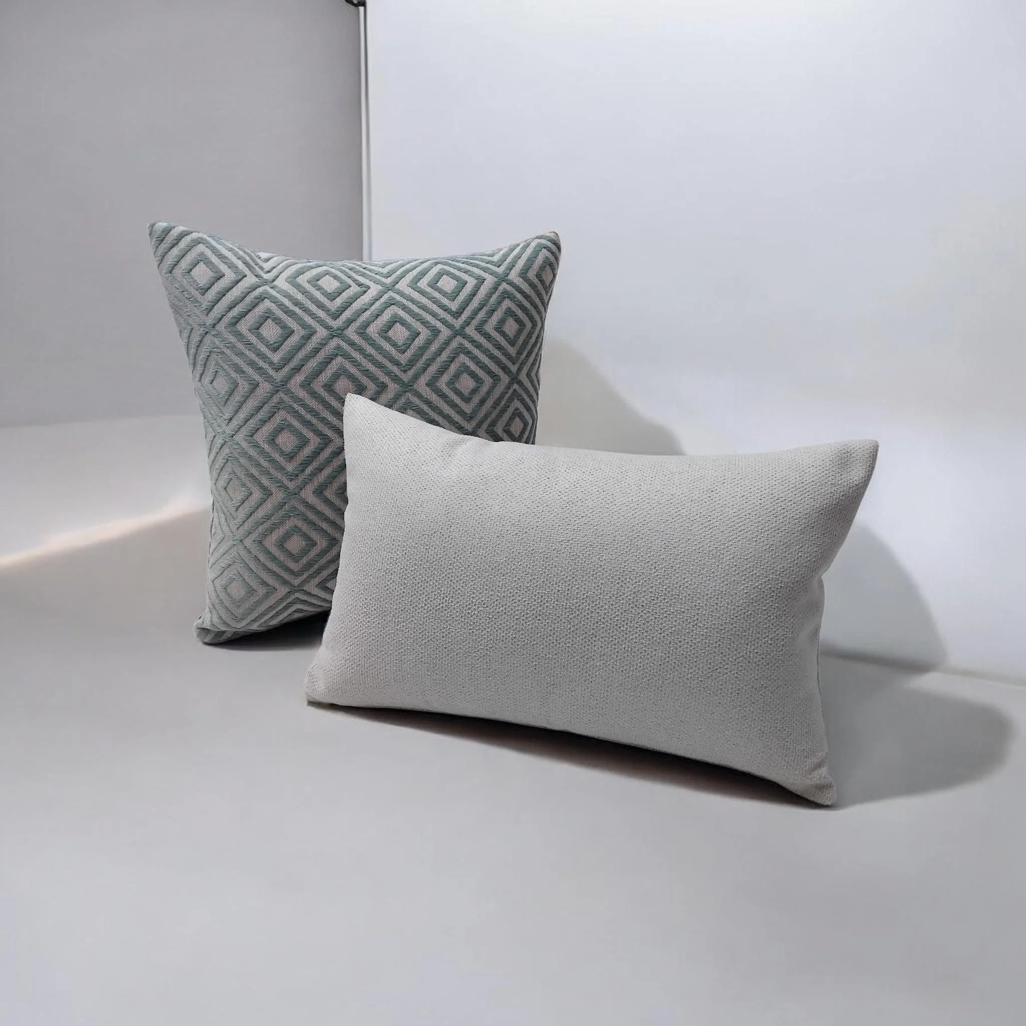 Close-up photo of two decorative pillows on a bed or sofa, one with a geometric diamond pattern in shades of gray and black, and the other in plain light gray fabric, against a plain white wall background.
