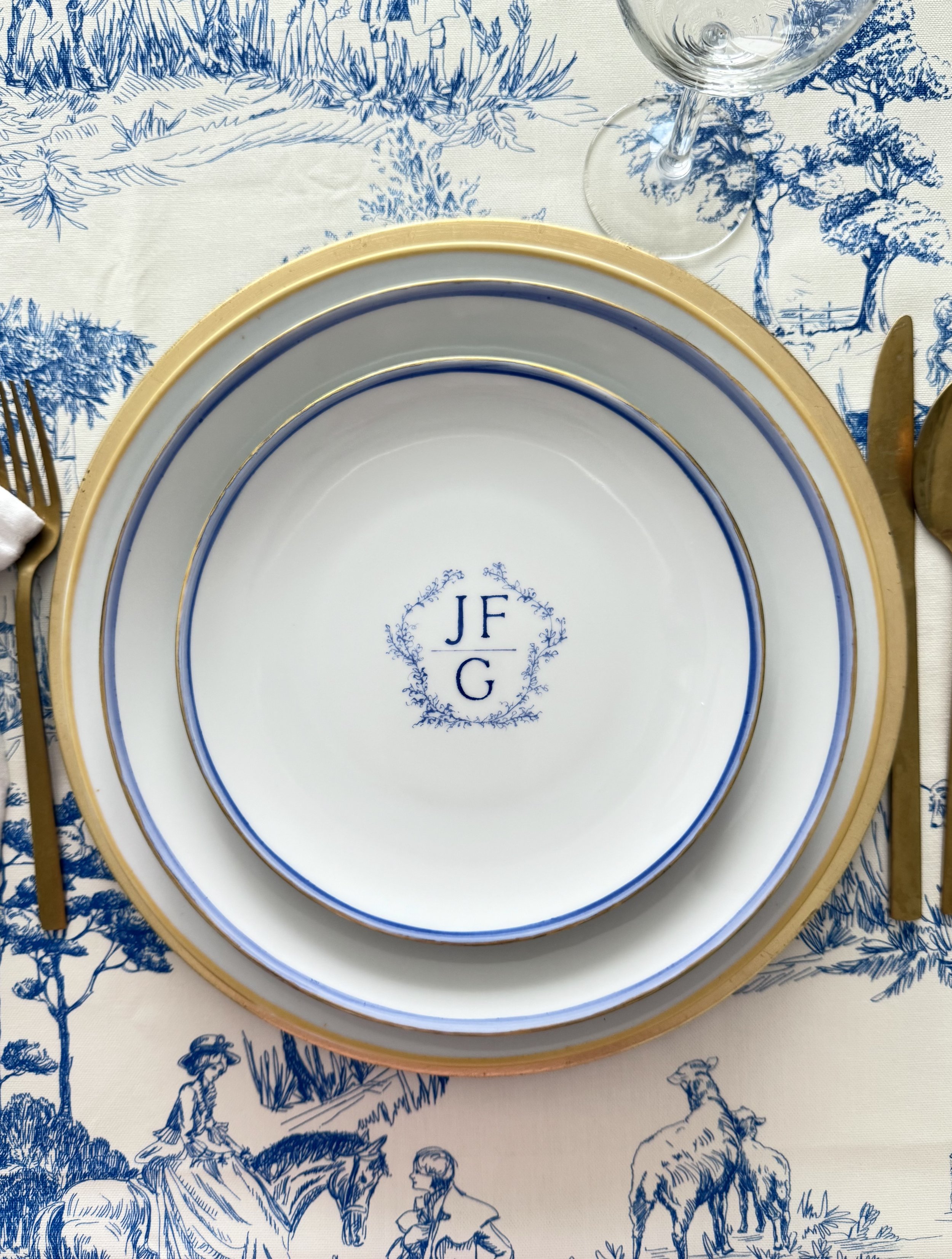 A set of white plates with blue and gold trim stacked on a table with a blue and white patterned tablecloth featuring pastoral scenes of people, trees, and animals. A clear wine glass is also visible.