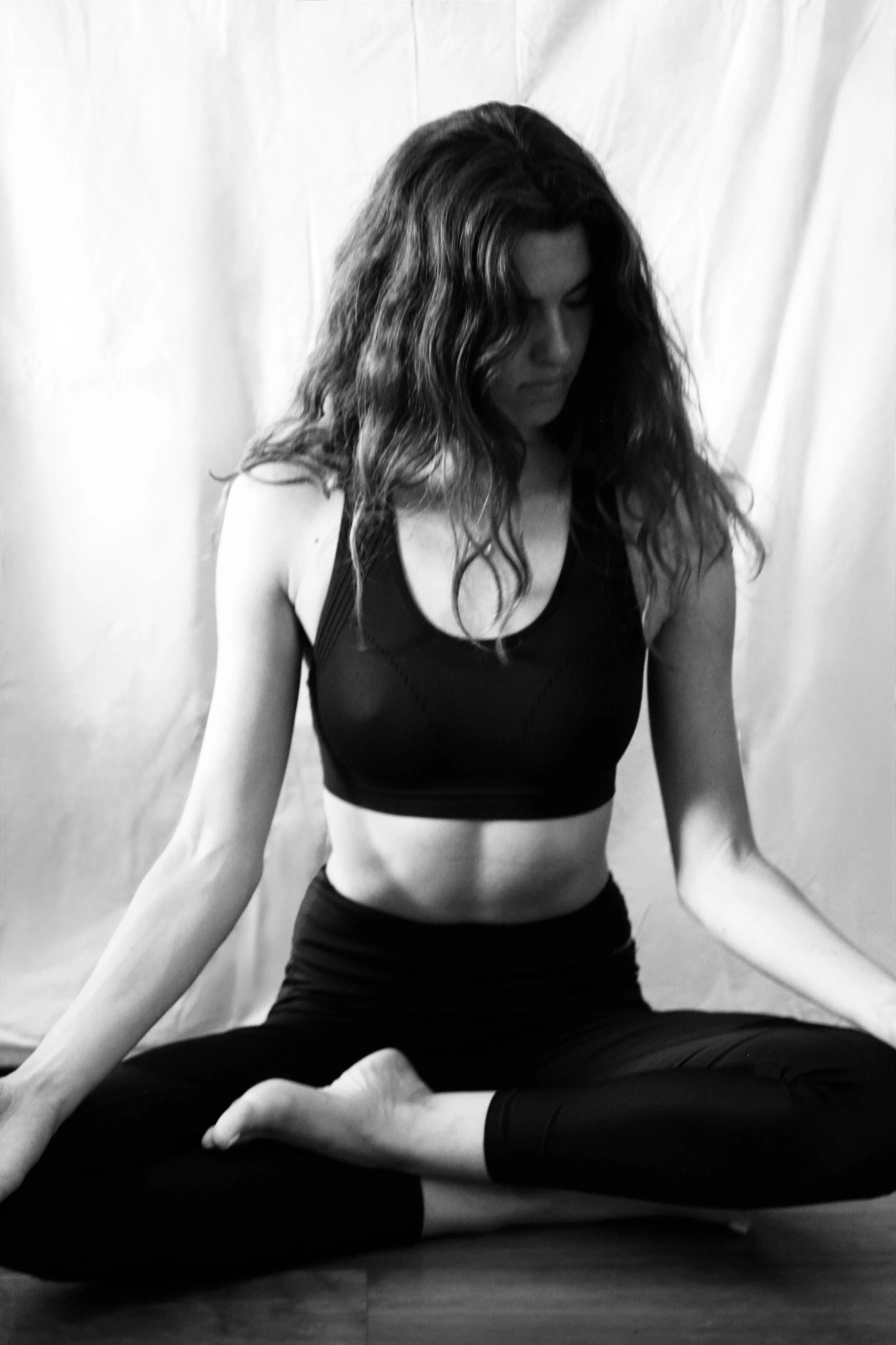 A woman practicing yoga in a seated pose on a wooden floor, with a white cloth background, wearing a sports bra and leggings.