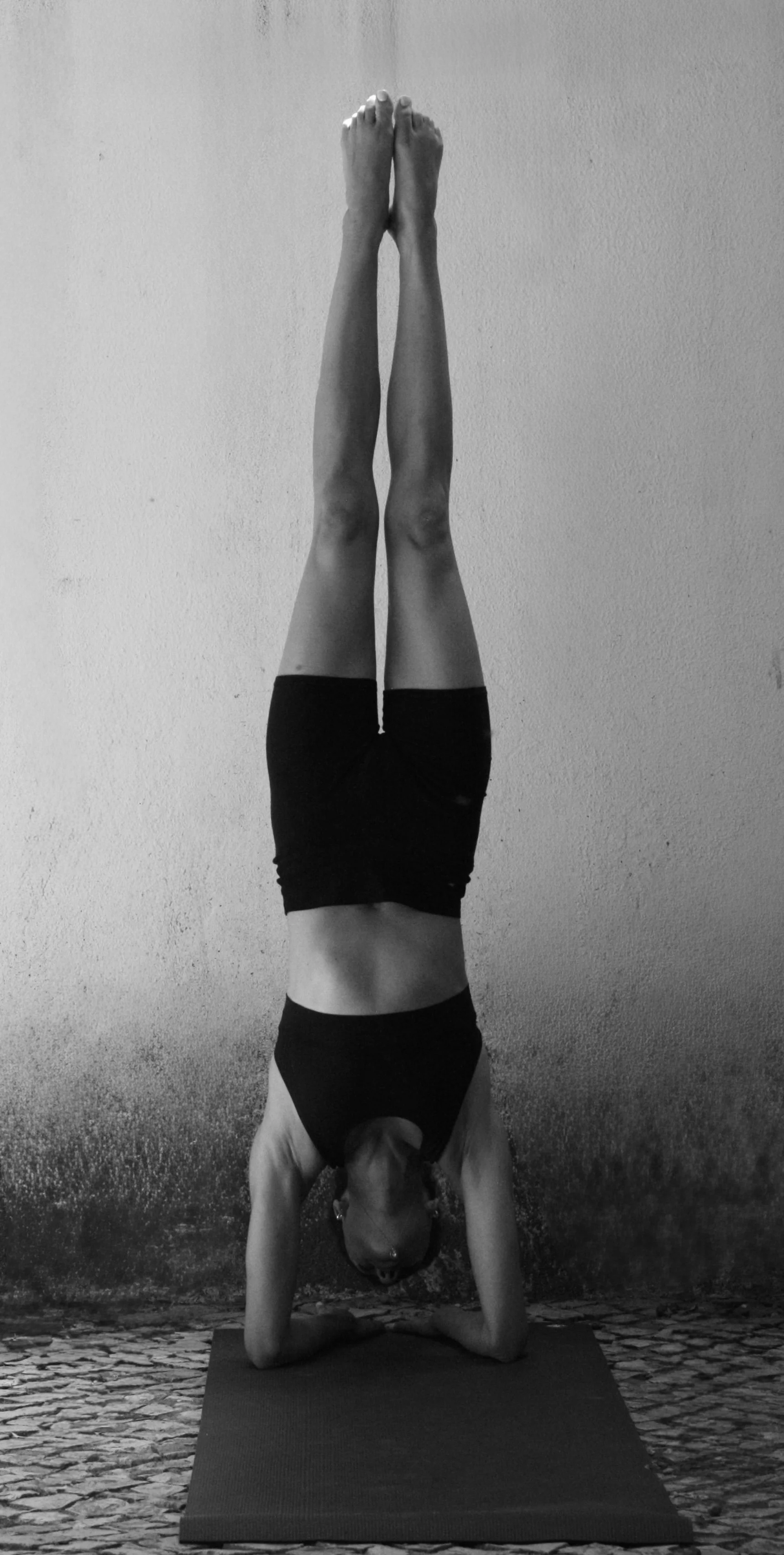 A person performing a headstand yoga pose on a yoga mat, wearing black shorts and a black sports bra, with one arm supporting their head and the other arm bent, against a plain textured wall.