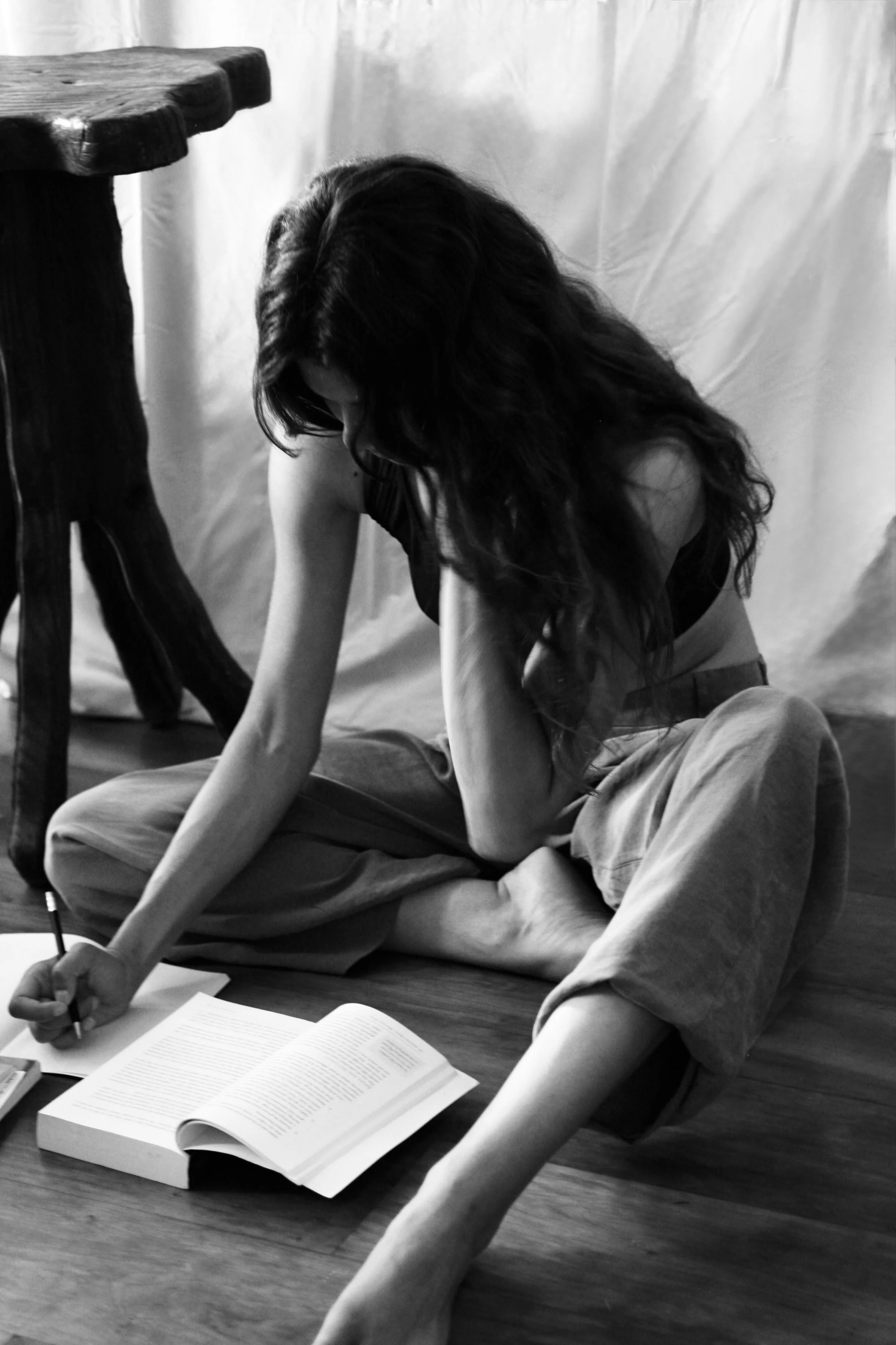 A woman with long dark hair is sitting on the floor, writing in a notebook, with an open book in front of her. There is a wooden table and a fabric backdrop in the background.