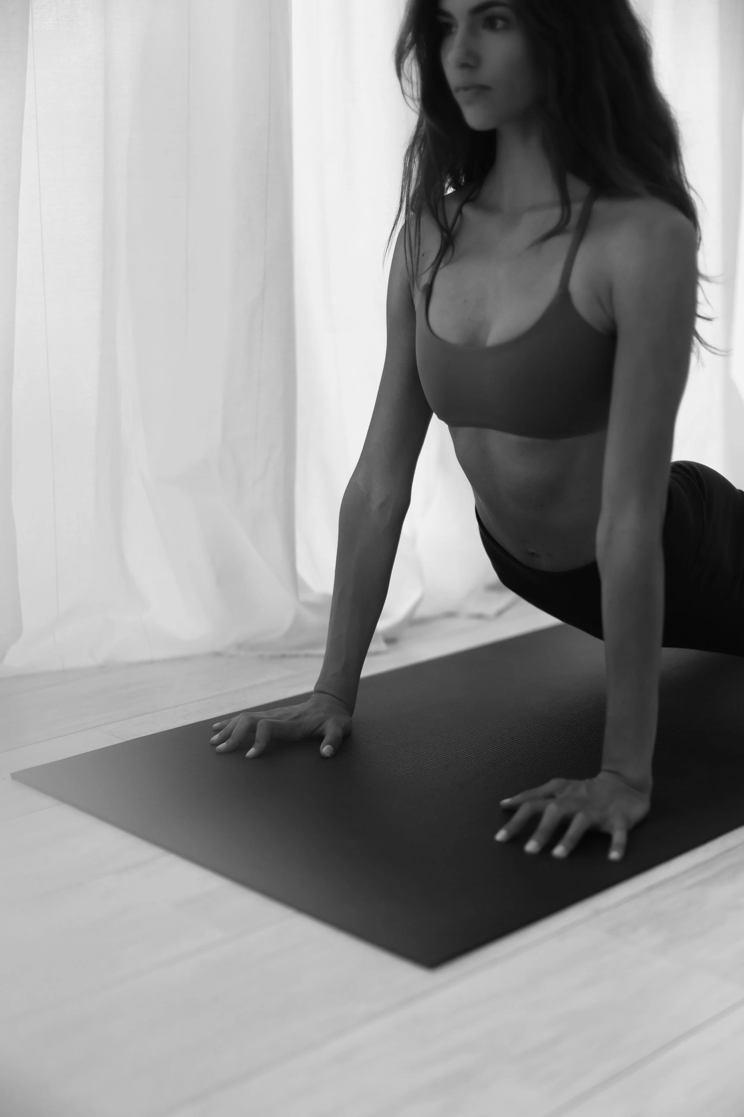 A woman in workout attire performing yoga or stretching on a yoga mat indoors, with light-colored curtains in the background.