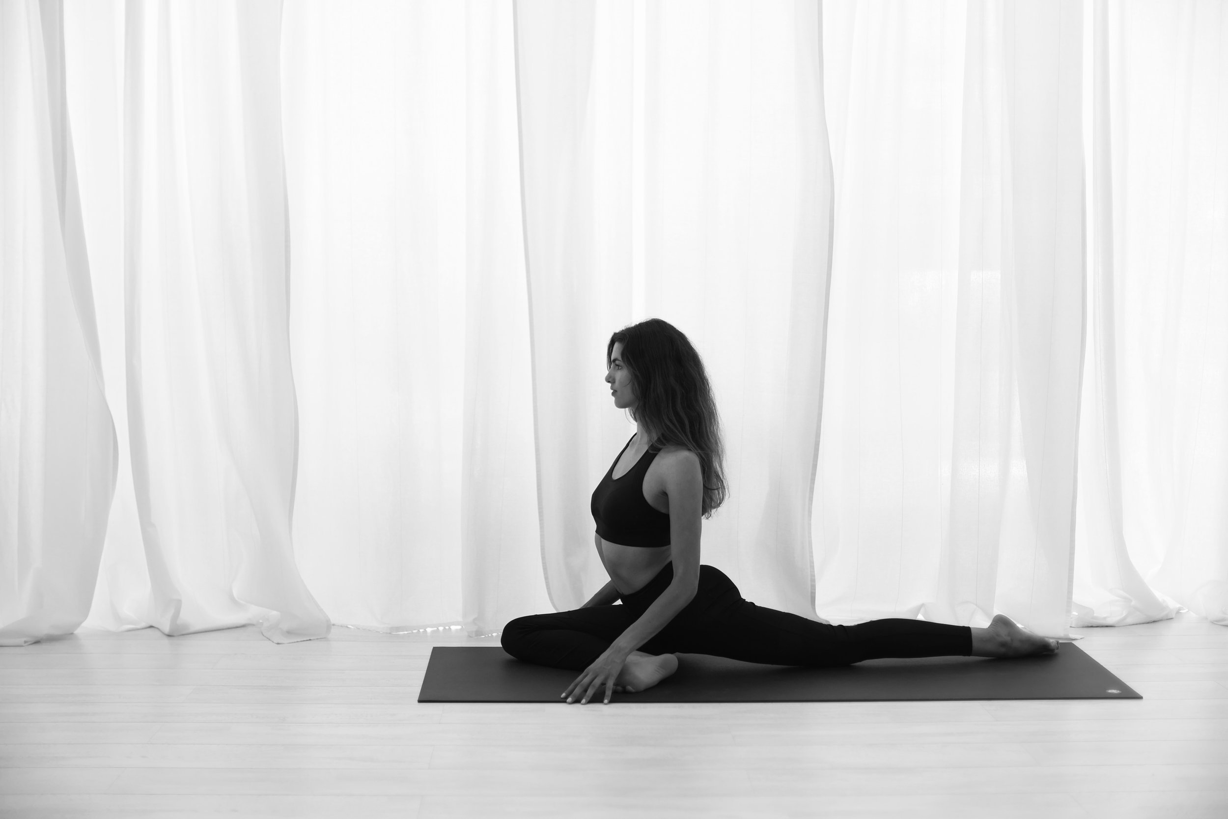 A woman practicing yoga in a black sports bra and leggings, performing a stretch on a yoga mat in front of a white curtain.