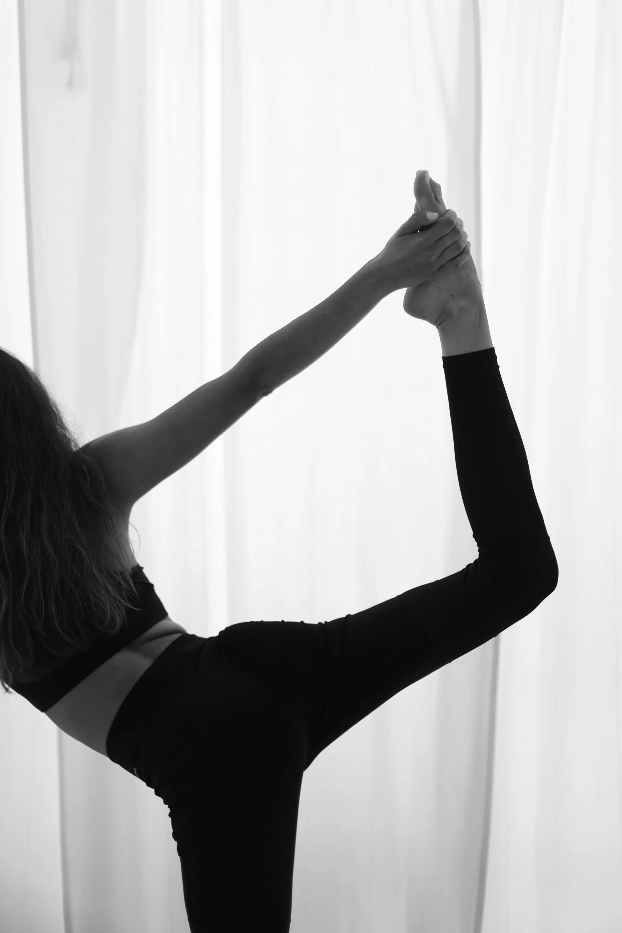 A person doing an advanced yoga pose with one hand holding the foot behind their head, in front of a light-colored curtain.