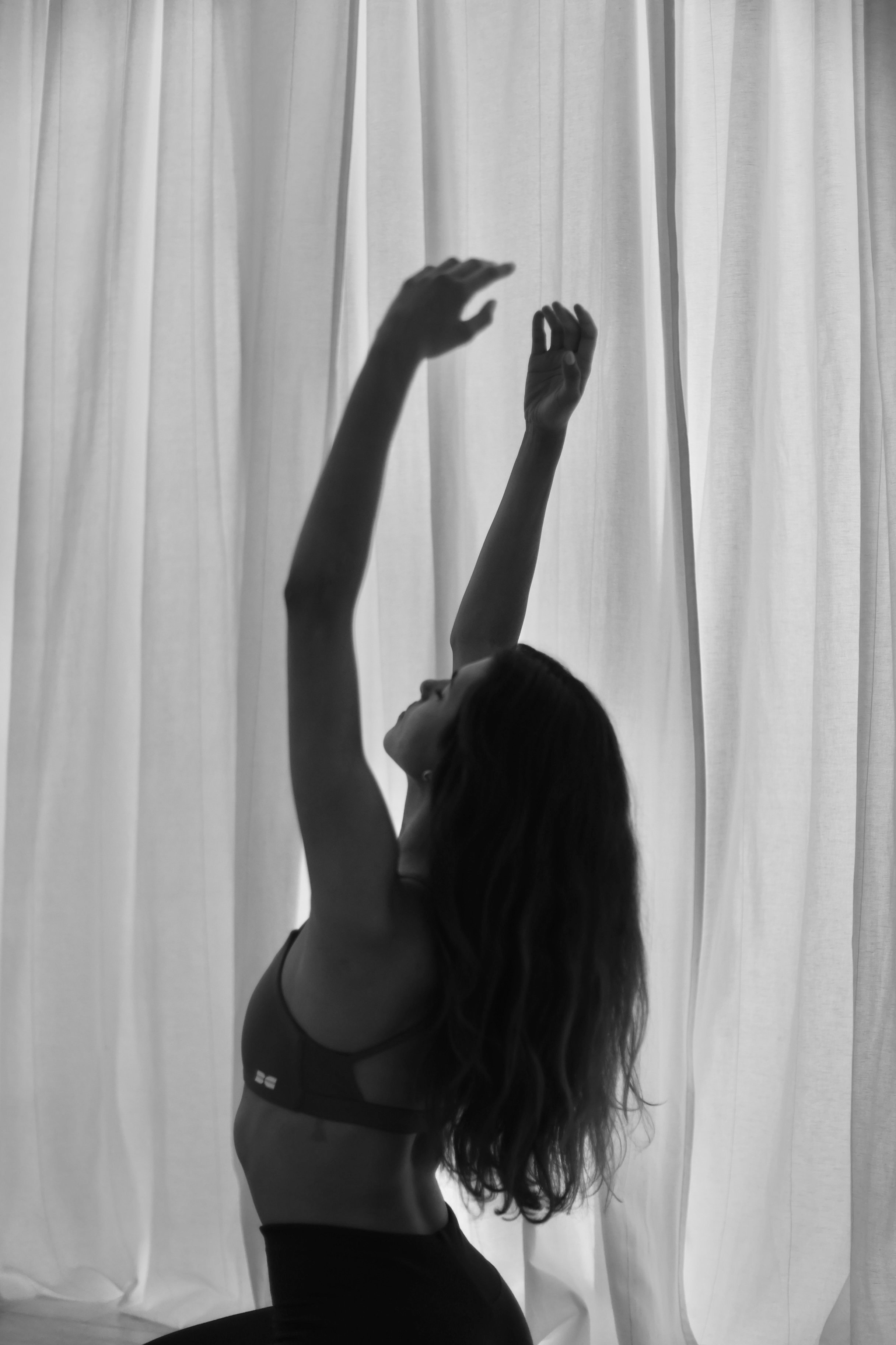 A black-and-white photo of a woman with long hair in a sports bra and leggings, stretching her arms upward in front of white curtains.