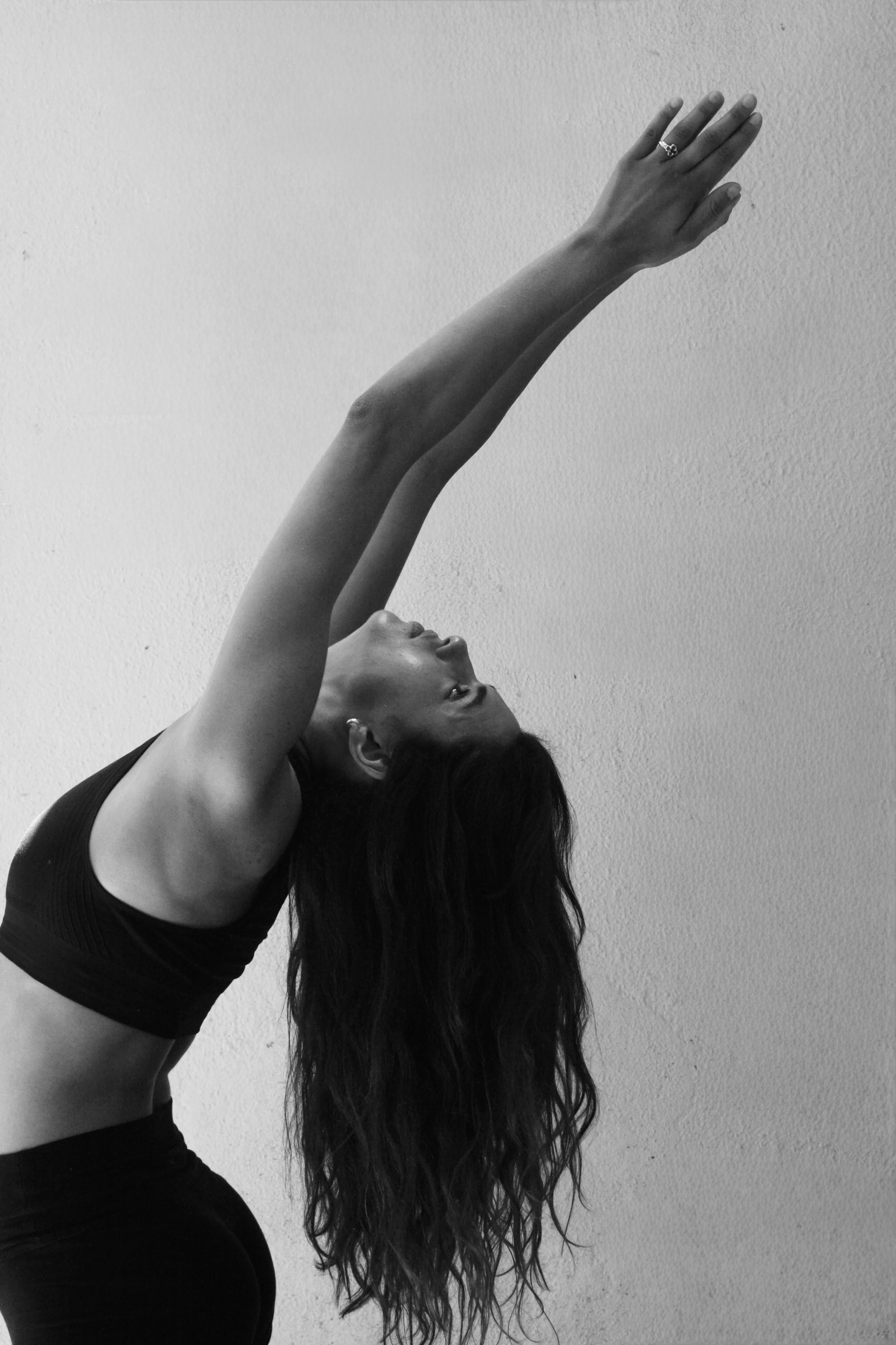 A woman wearing a black sports bra stretching her arms above her head, with her head tilted back and long hair hanging down.