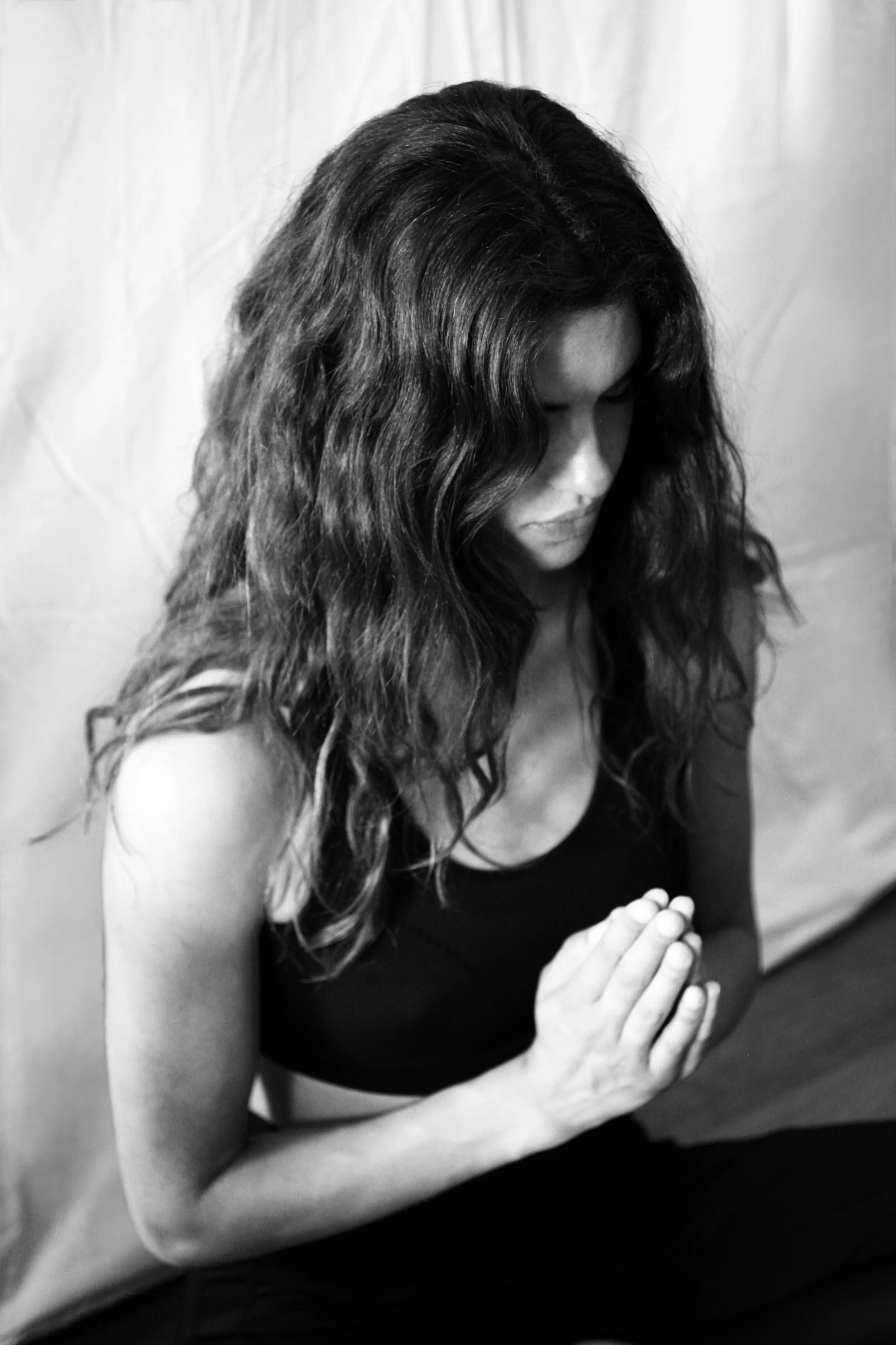A woman with long, wavy hair is in a prayer or meditation pose with her eyes closed and hands pressed together in front of her chest.