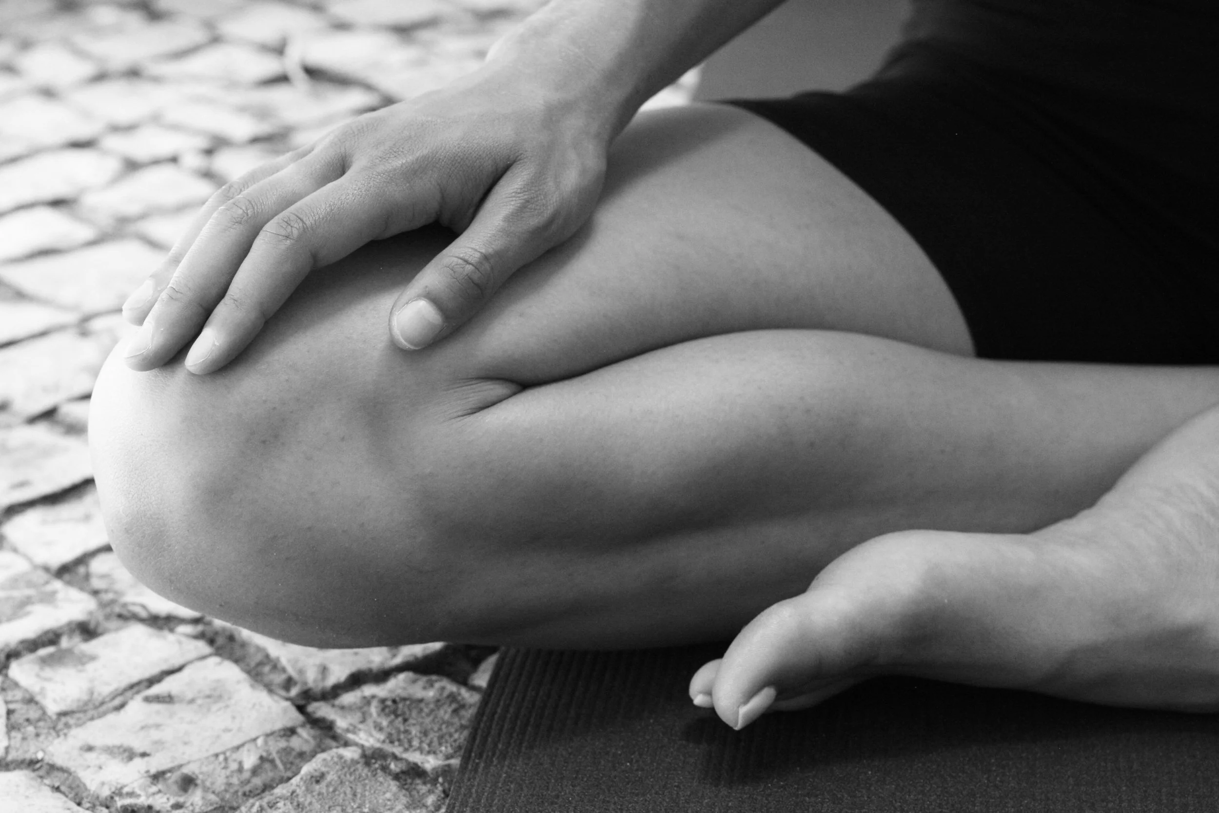 Black and white close-up photo of two hands clasped on a person's knee, with textured stone pavement in the background.