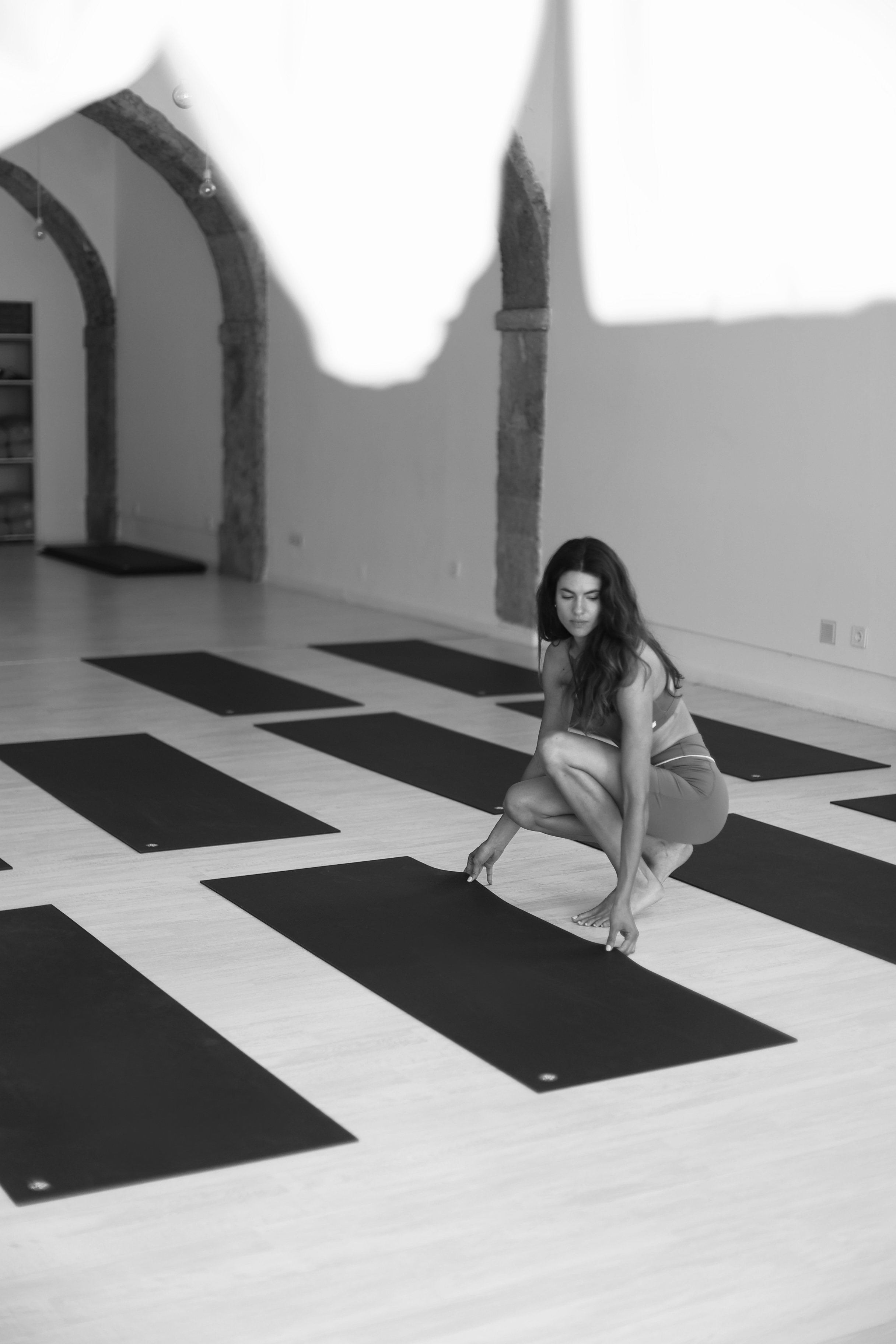 A woman practicing yoga on a mat in a spacious room with arched stone columns, with her hand reaching towards her foot.