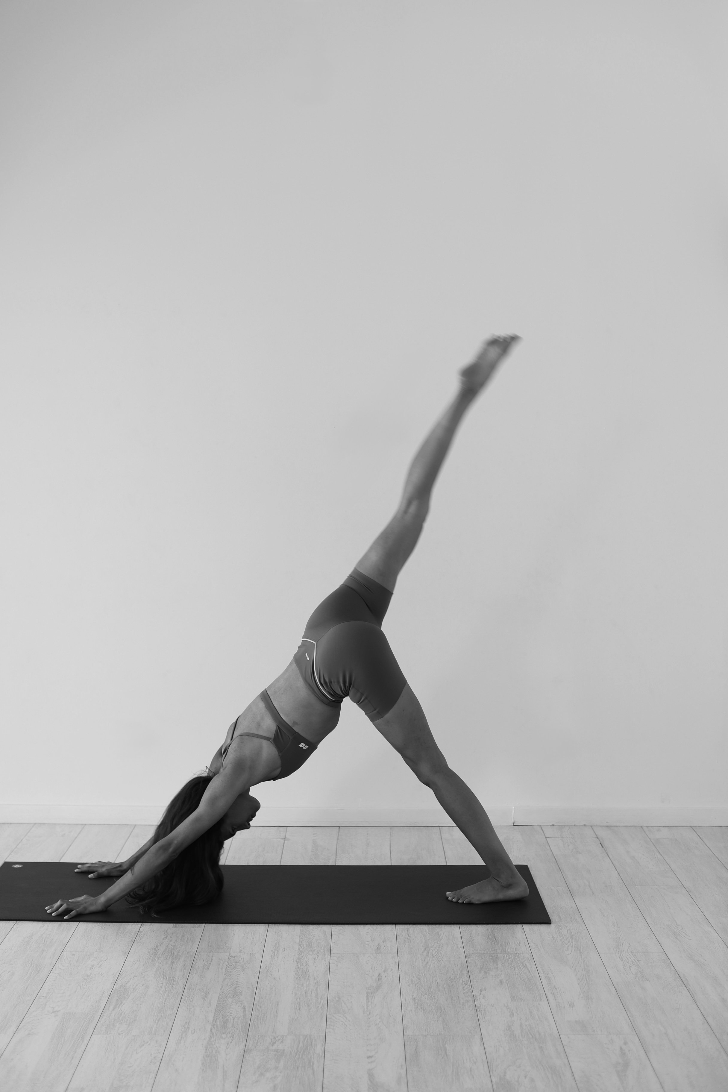 A woman practicing yoga indoors, performing a handstand with her legs extended upwards and slightly bent, on a yoga mat against a plain wall.
