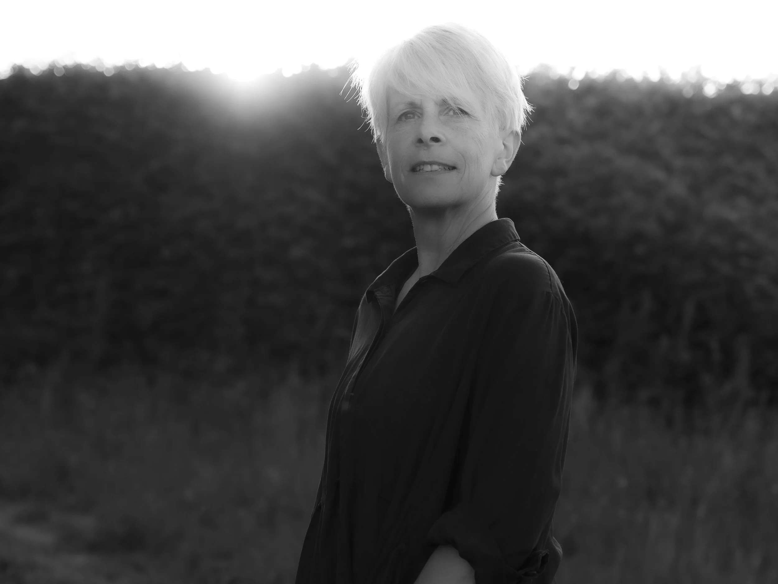 A black-and-white photograph of an older woman with short, light hair outdoors during sunset or sunrise, looking over her shoulder with a thoughtful expression, with blurred natural landscape in the background.