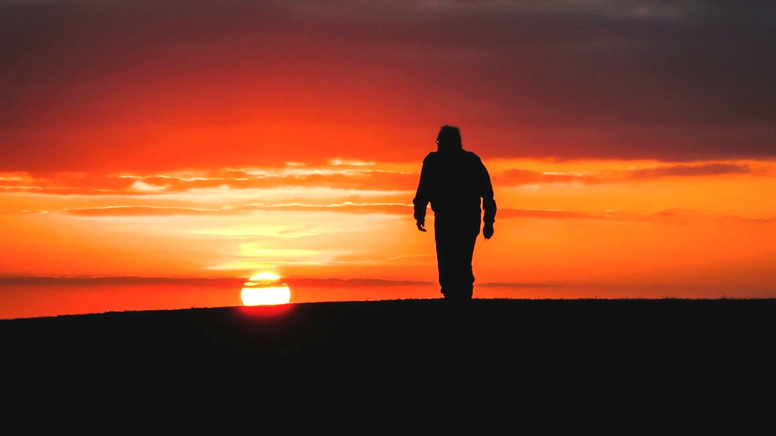 Person walking along the horizon during a vibrant sunset with orange, red, and purple sky.
