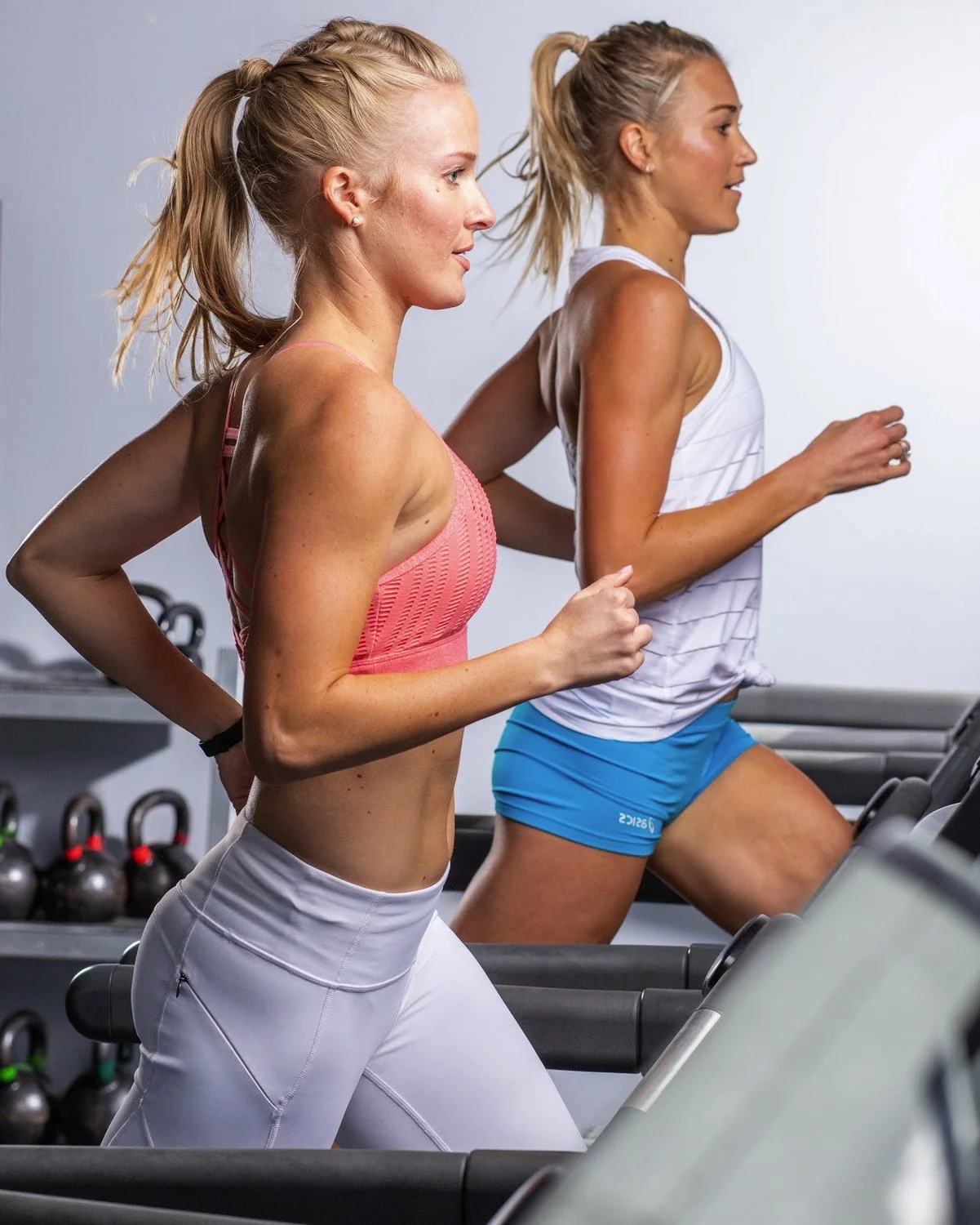 Two women jogging on treadmills in a gym. One wears a pink sports bra and white leggings, the other a white top and blue shorts.