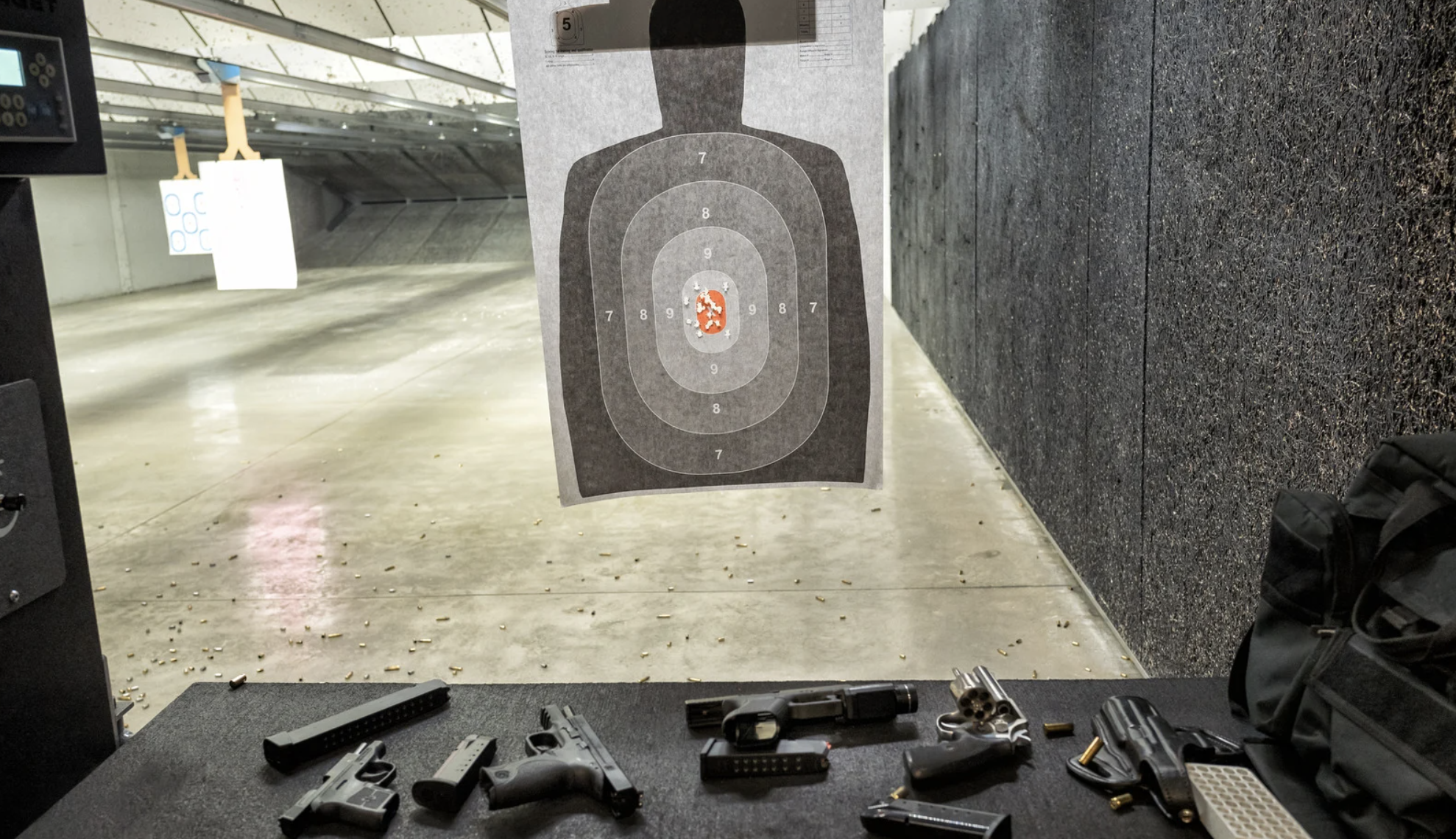 Indoor shooting range with a paper target featuring a silhouette of a person, surrounded by concentric scoring rings, with spent shell casings scattered on the floor and pistol firearms on a table in the foreground.