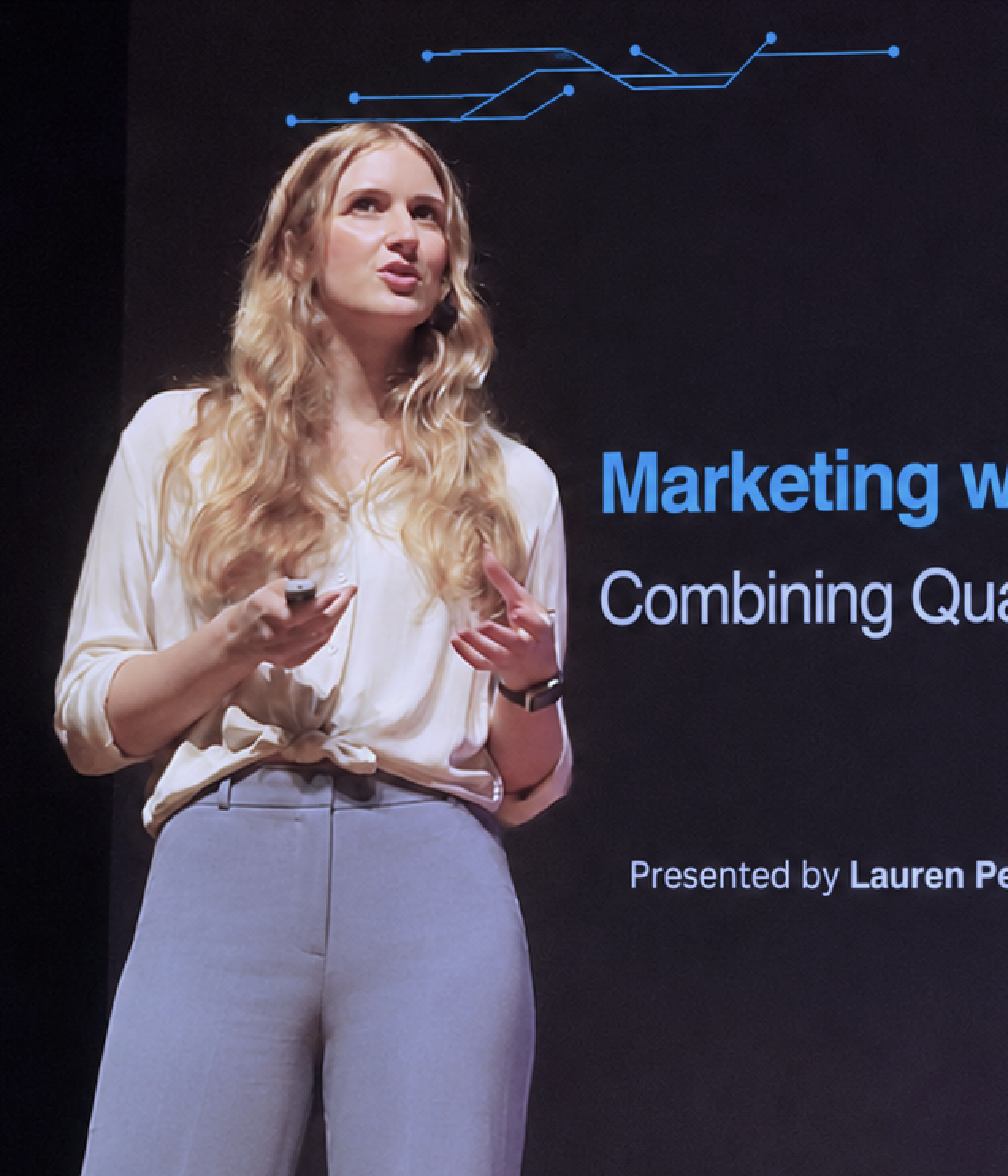 A woman with long wavy blonde hair giving a presentation on marketing, standing in front of a black screen with blue and white text.