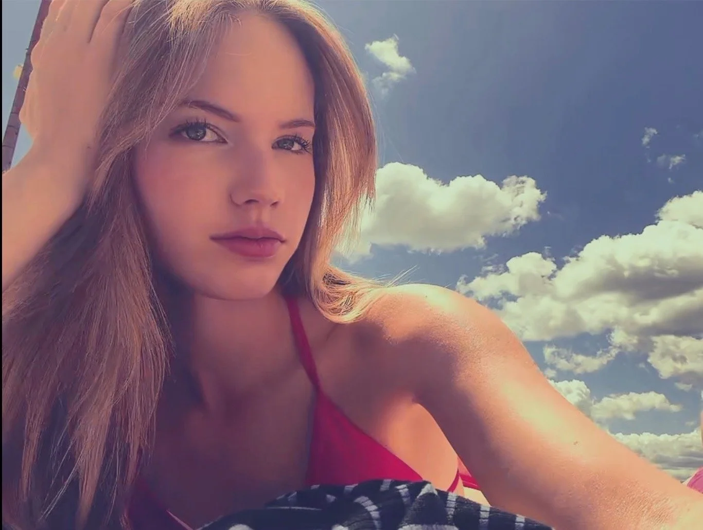 A woman with long reddish-brown hair, wearing a red top, posing outdoors on a sunny day with a blue sky and white clouds in the background.
