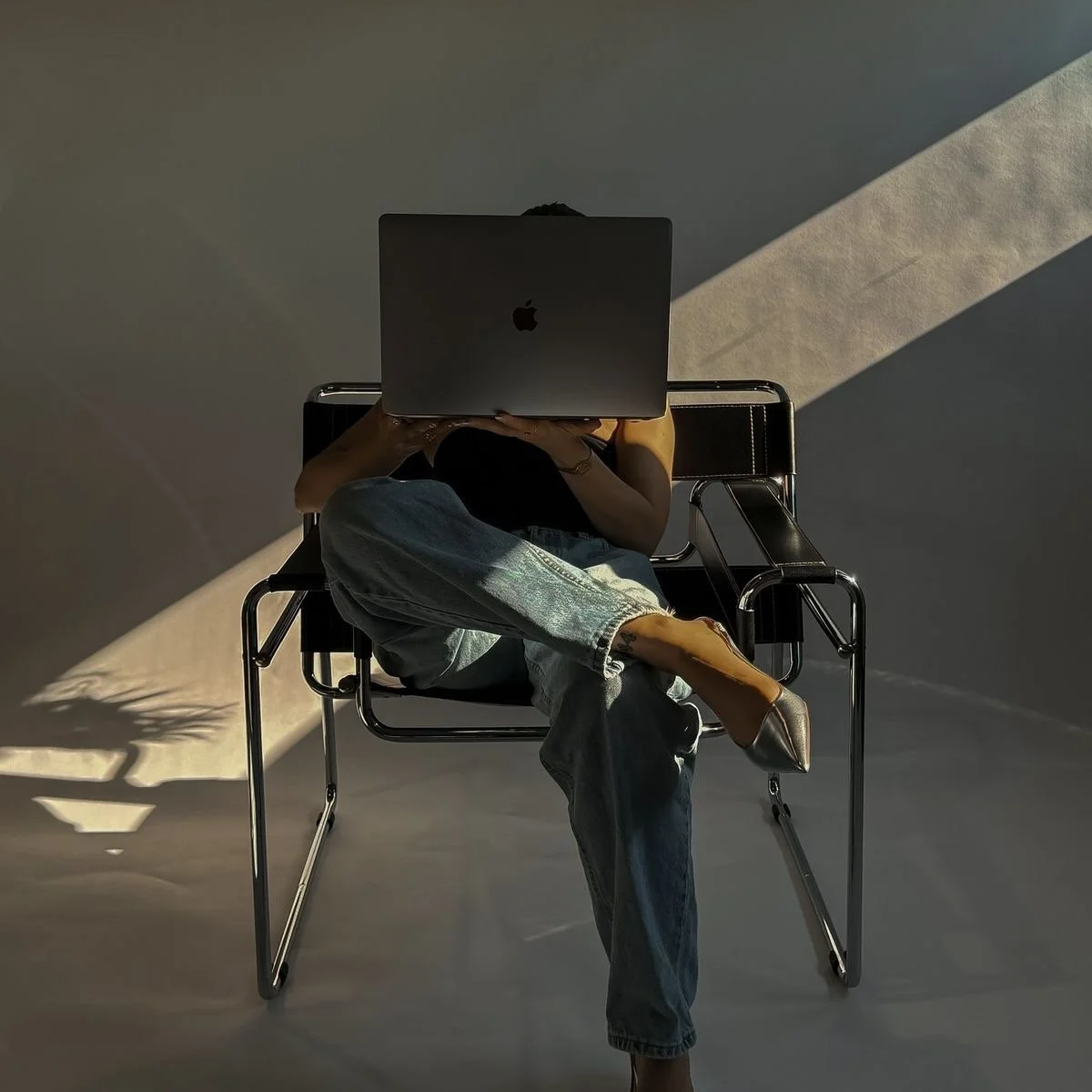 Person sitting on a black chair, using a laptop, with sunlight casting shadows on the wall behind.
