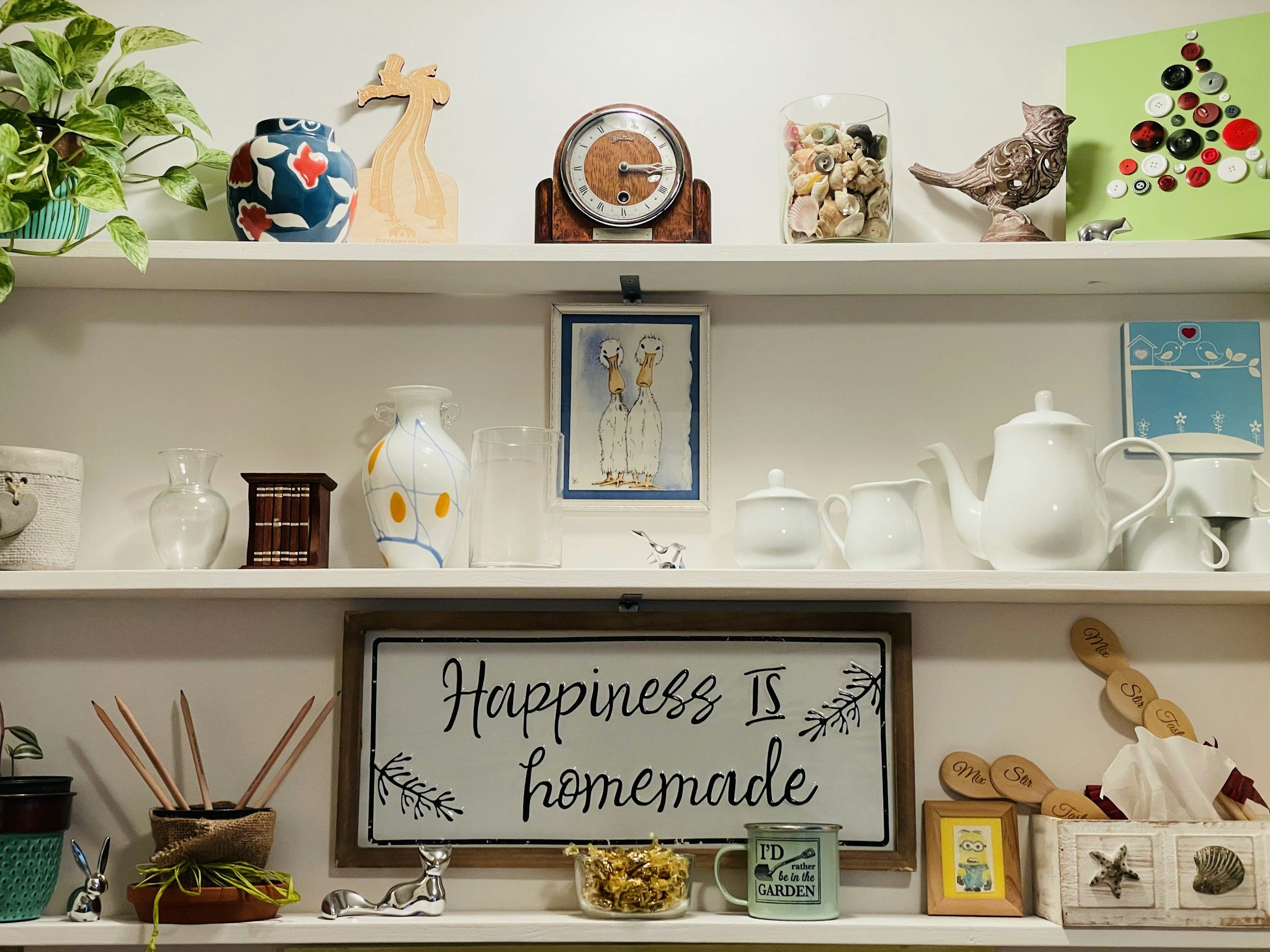 Decorative shelf with a potted plant, ceramic vase, wooden giraffe, clock, glass jar filled with shells, ceramic bird sculpture, green board with buttons, drawers, various white ceramic dishware, framed illustration of two birds, and a white sign with black lettering.