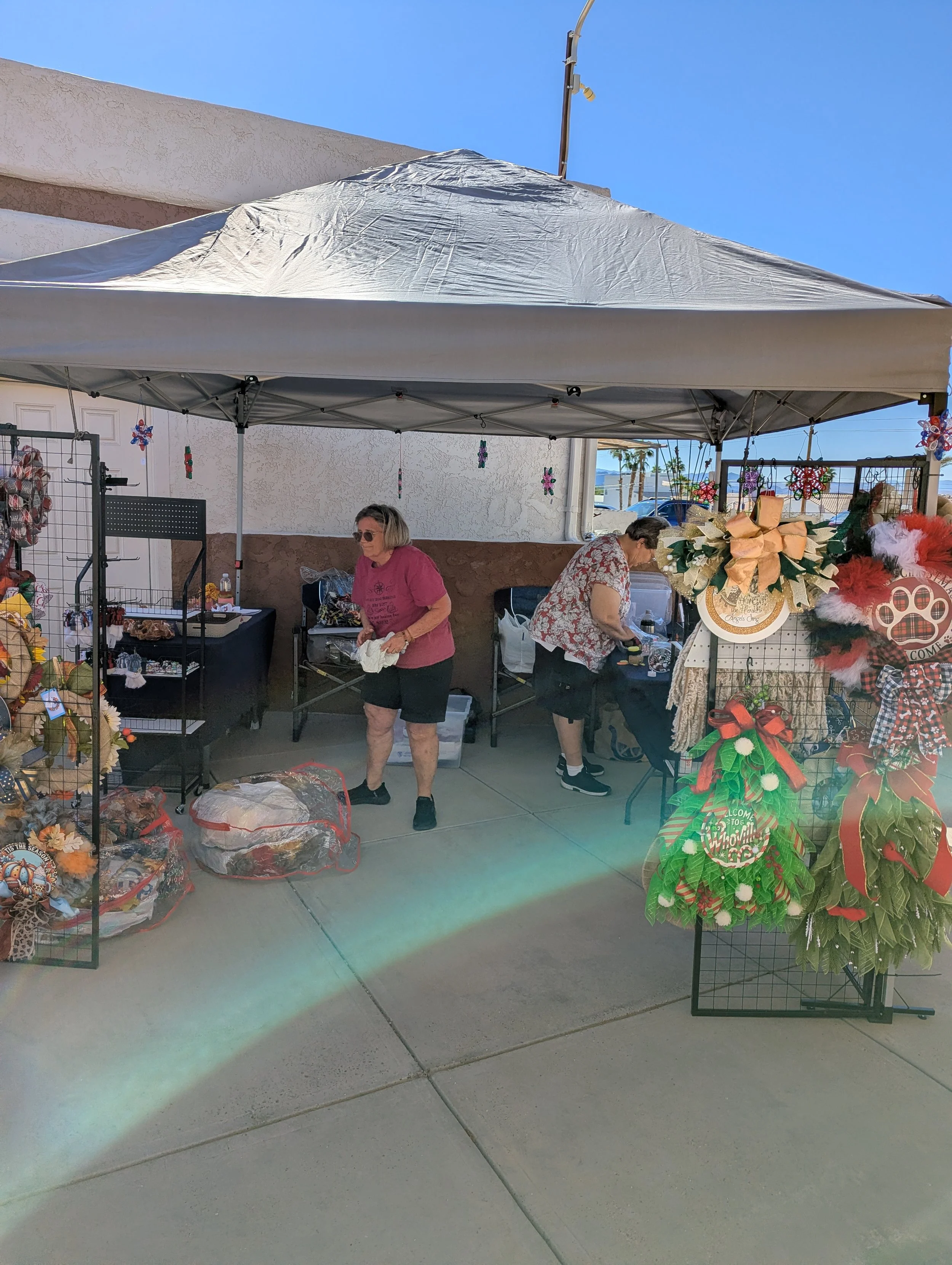 Outdoor market stall with Christmas decorations, including poinsettias and bows, under a white canopy and against a beige wall with a clear blue sky in the background.