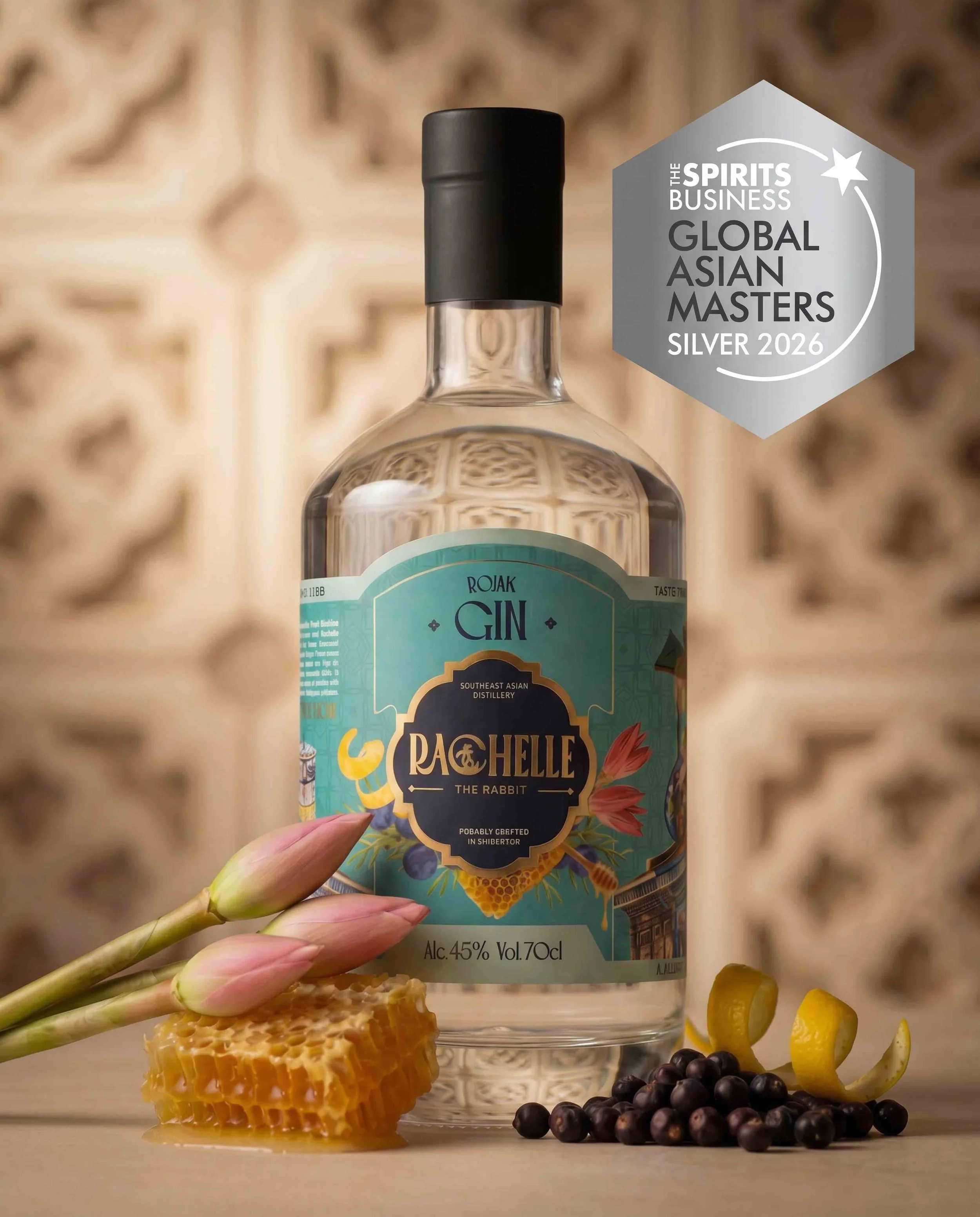 A hand holding a bottle of Southeast Asian gin labeled 'Rachel the Rabbit' with a honeycomb, blackcurrants, and lemon slices arranged on a surface in front of it. A blurred ornate background is also visible.