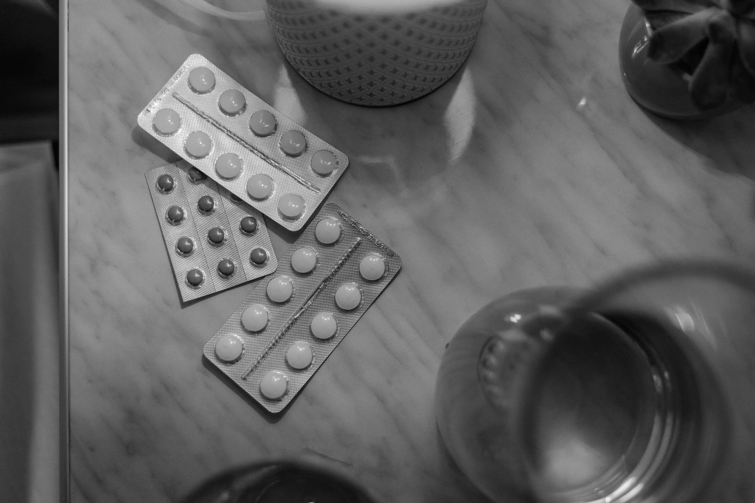 Black and white photograph of a table with several blister packs of pills scattered across, a small metal object, and two glass jars, one with a textured pattern and the other with a reflective surface.