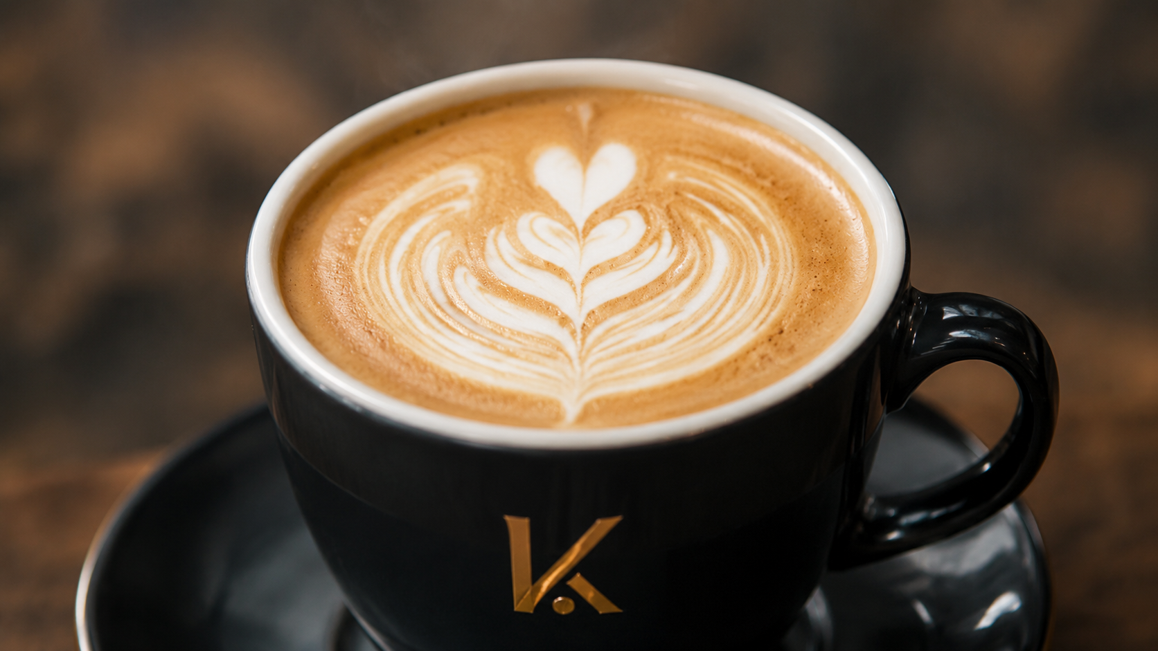 Taza de café con arte en leche en una taza negra con logo dorado, sobre un plato negro.