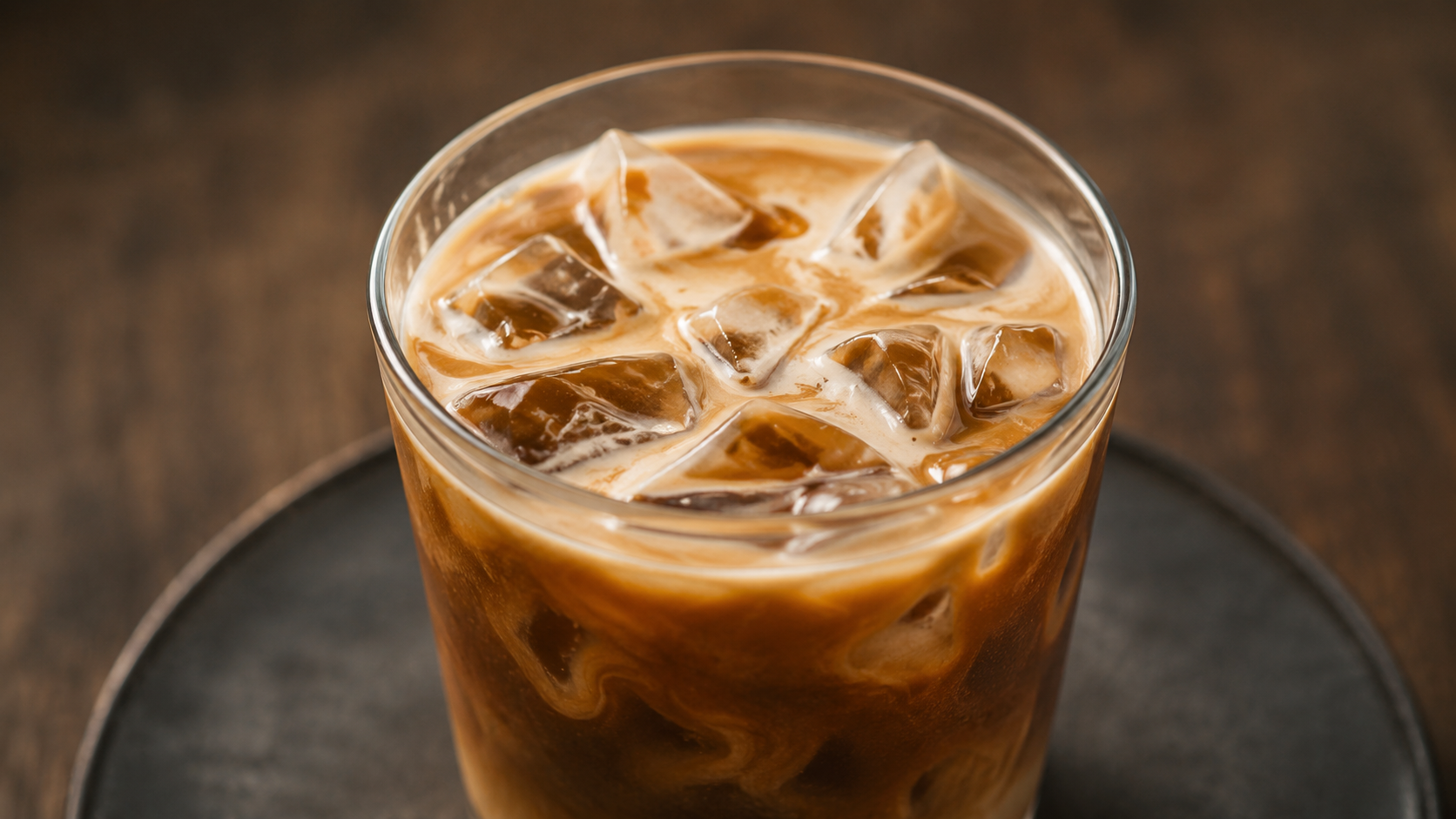 Vaso de vidrio con hielo y café con leche, visto desde arriba sobre una bandeja negra.