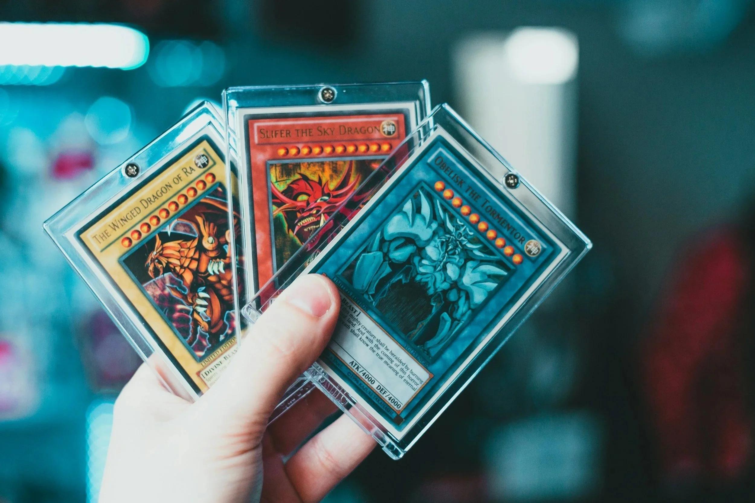 Hand holding a small sleeve containing three Yu-Gi-Oh! trading cards, with colorful blurred background.