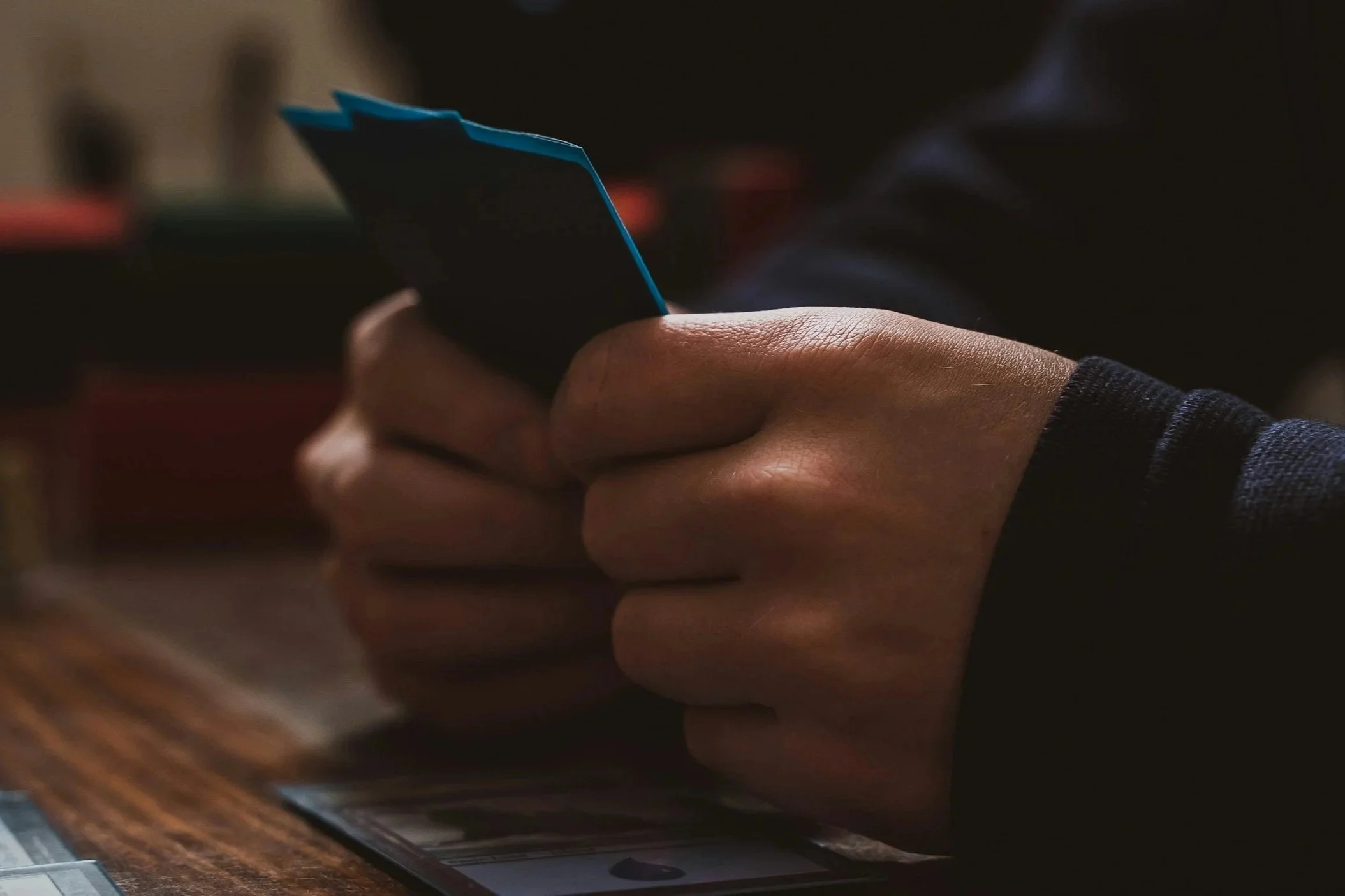 Close-up of a person's hand holding a blue card, with a blurred background, dark lighting, and a wooden surface visible at the bottom.