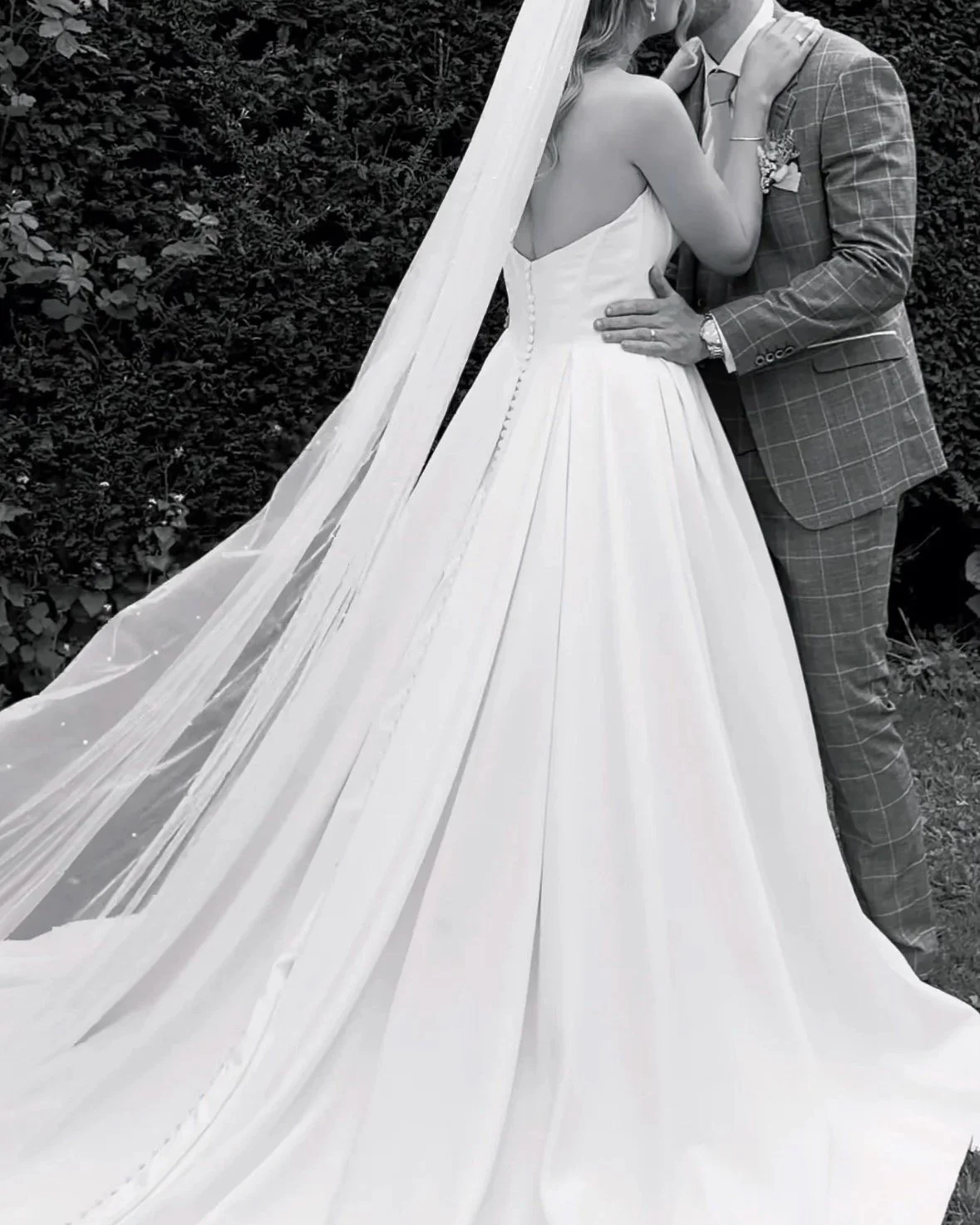 A bride and groom embracing on their wedding day, with the bride wearing a strapless white gown and the groom in a checked suit, standing outdoors with a leafy background.