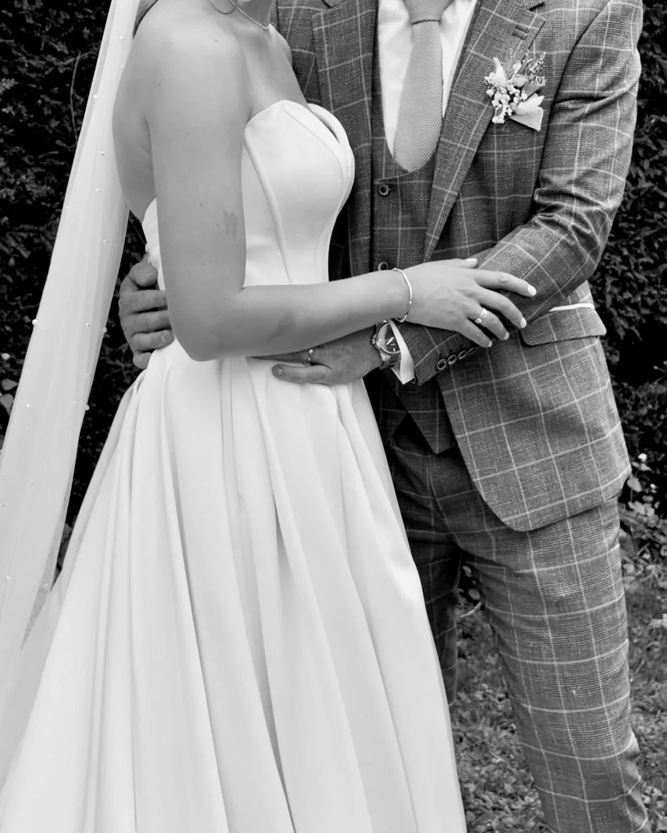 A bride and groom holding hands in wedding attire, the bride in a strapless white wedding gown and the groom in a plaid suit with a boutonniere, outdoors in black and white.